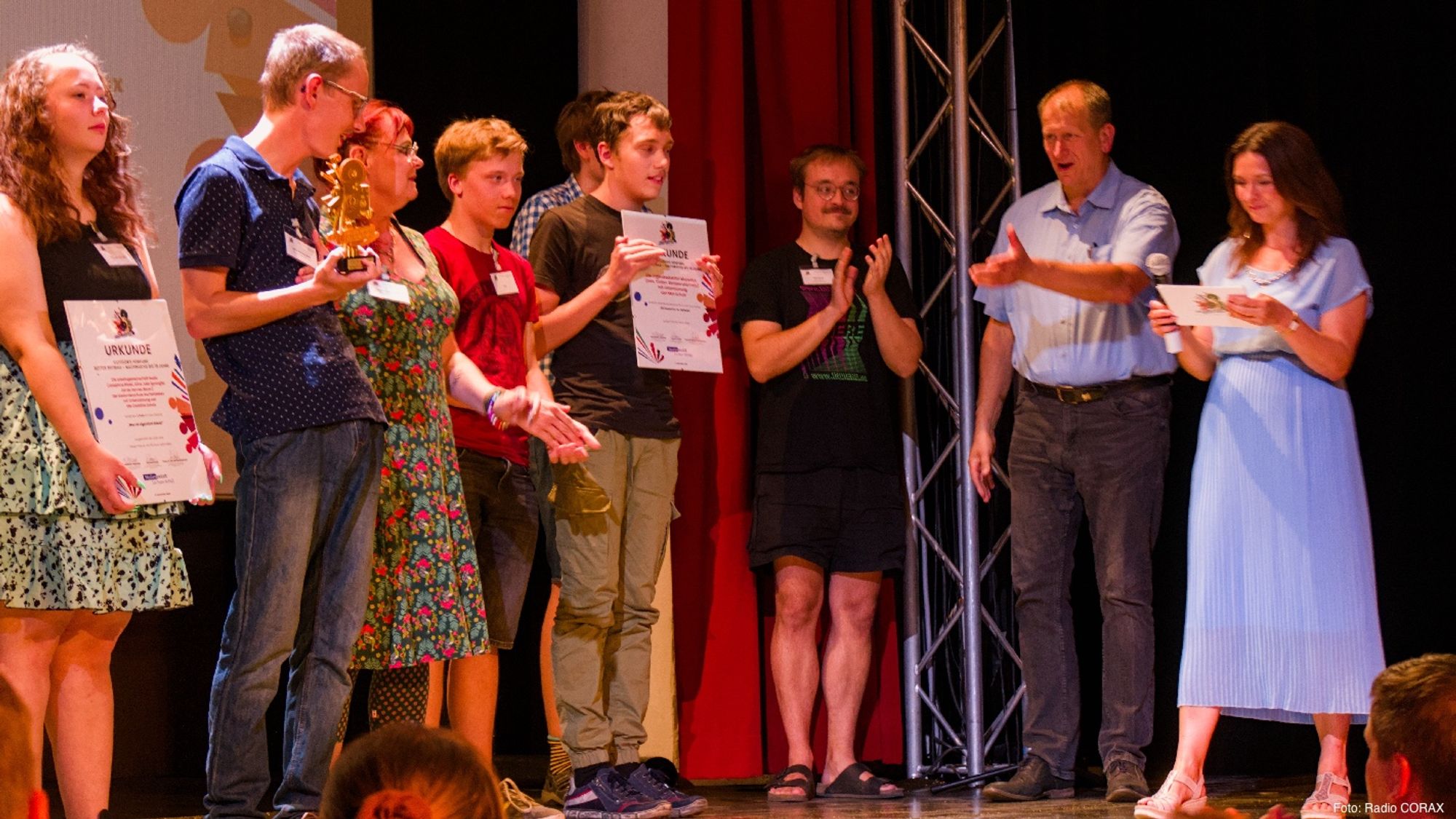 Das Bild zeigt viele Menschen auf einer Bühne, davor ist das Publikum erkennbar. Ganz rechts steht eine Frau im blauen Kleid, die auf eine Moderationskarte schaut und ein Mikrofon in der Hand hält. Neben ihr steht ein Mann mit blauem Hemd, der würdigend seine Hand hebt. Von den anderen, hauptsächlich jungen Menschen halten zwei eine Urkunde und einer einen Preis in der Hand, die anderen klatschen.