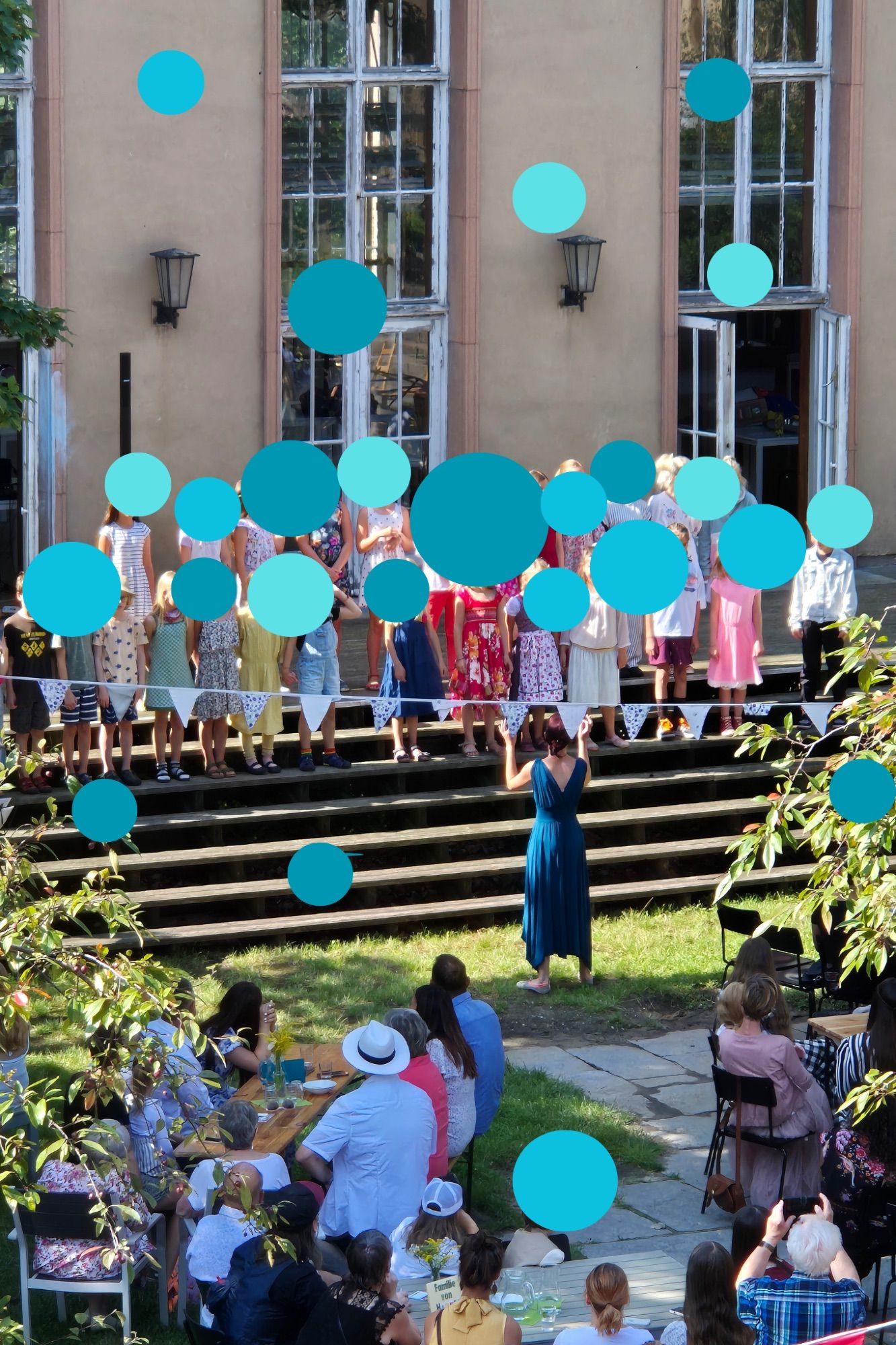 Auf den Terrassentreppen vor dem Haus stehen viele unkenntlich gemachte Kinder als Chor, davor eine Dirigentin und hinter ihr viele Gäste an Tischen unter einen Wimpelkette.