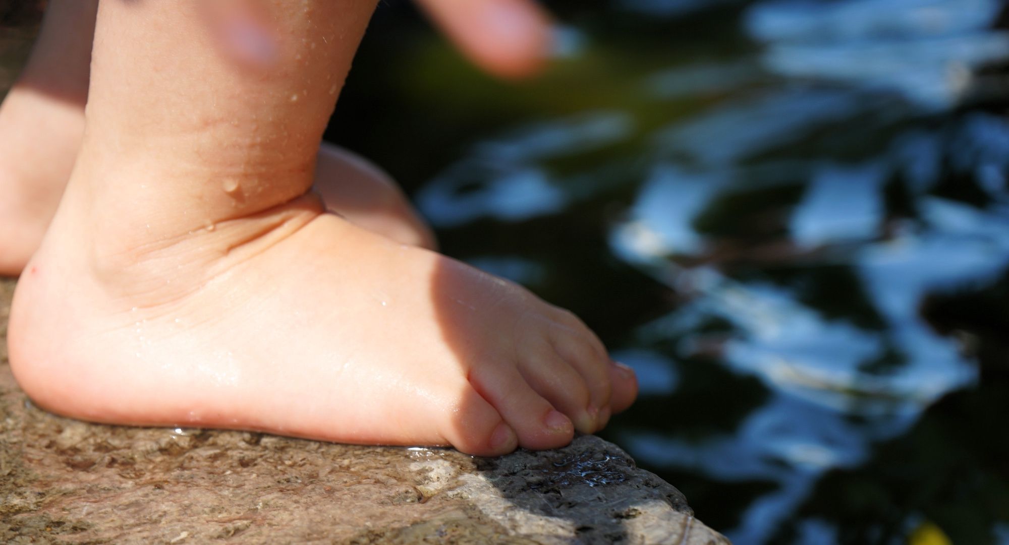 Das Bild zeigt eine Nahaufnahme eines nassen Kinderfußes, der auf einem Stein oder einer Art Beckenrand steht, daneben ist Wasser.