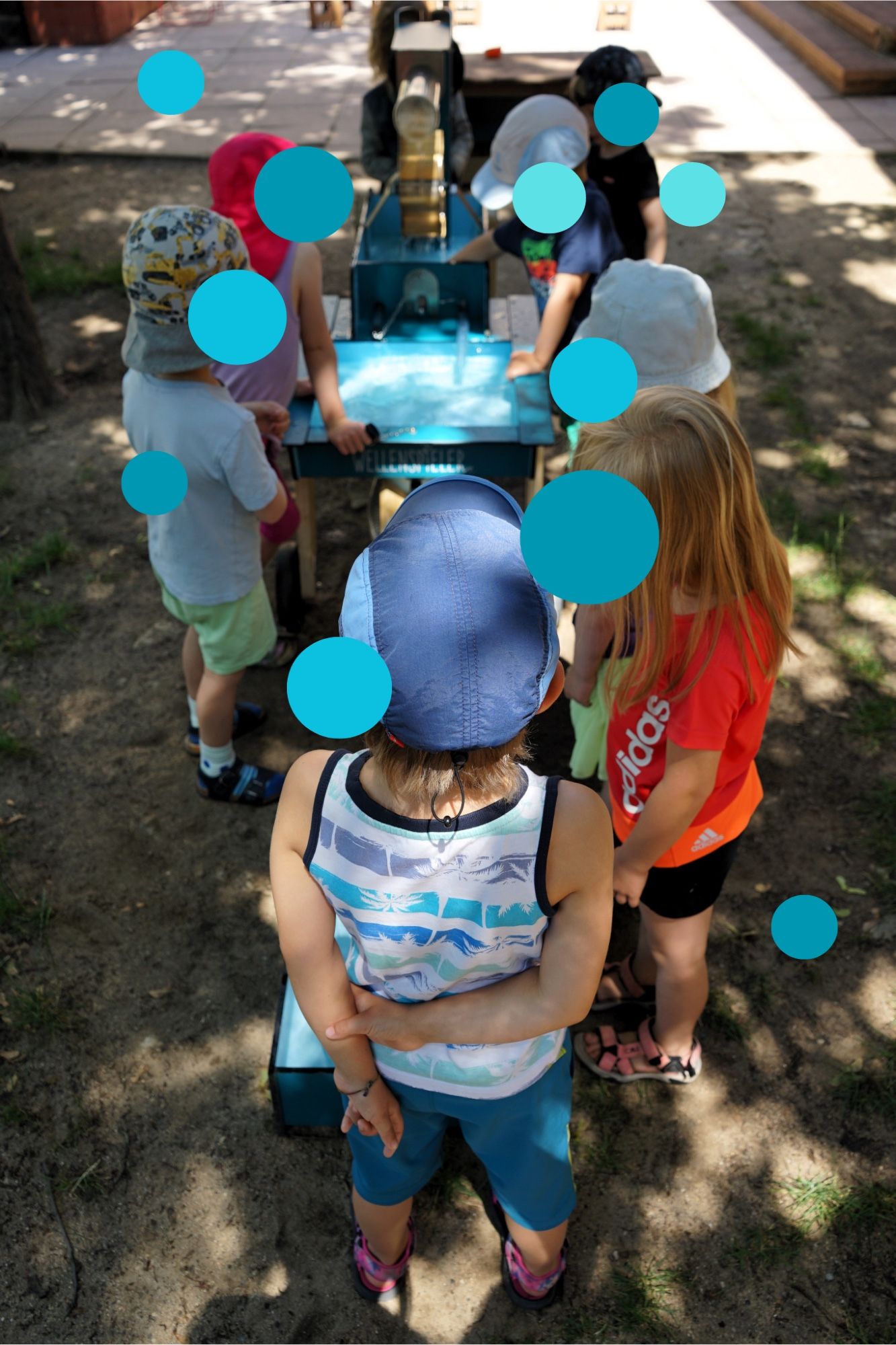 Das Bild zeigt sieben mit blauen Konfettipunkten unkenntlich gemachte Kinder in Sommerkleidung um einen Wellenspieler stehen.