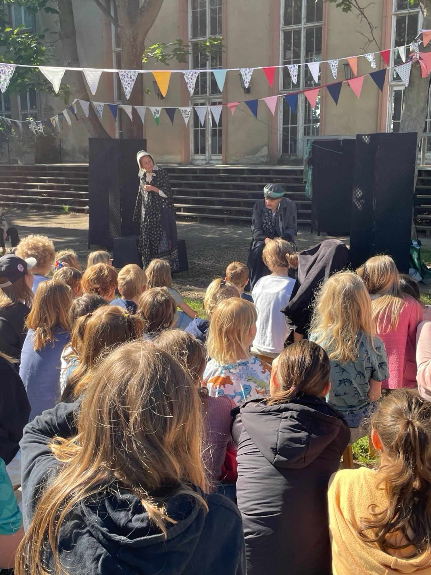 Auch hier sind viele Kinder von hinten zu sehen, die auf die Bühne mit farbenfrohen Wimpeln und schwarzen Requisiten blicken: der Schauspieler mit grauer Jacke und Mütze sitzt. Die Schauspielerin steht daneben im historischen dunklen Gewand mit weißer Haube.