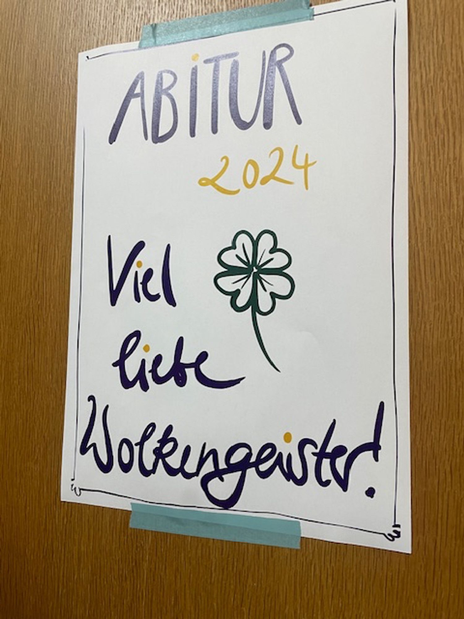 Das Bild zeigt ein Schild an einer Tür, auf dem in schönen blauen und gelben Buchstaben mit einem vierblättrigen Kleeblatt „Abitur 2024“ steht.