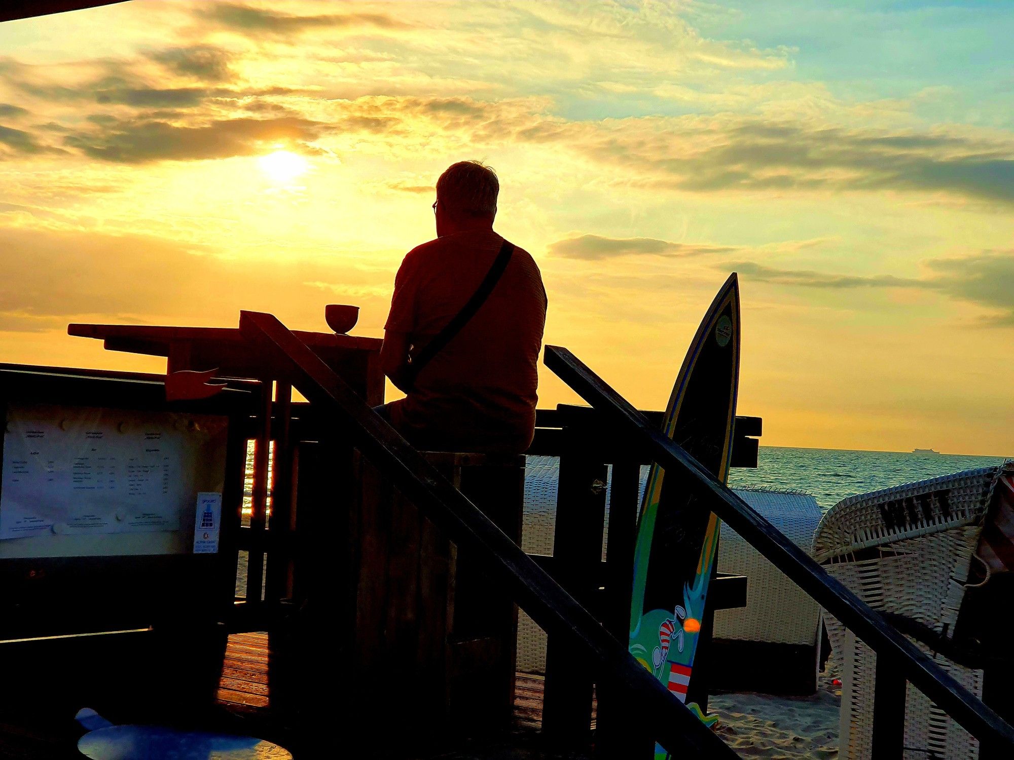 Sonnenuntergang in Goldgelb an der Ostsee. Gegenlichtaufnahme. Im Vodergrund als schwarze Silhouette ein Mensch an einem Bistrotisch auf einer kleinen Plattform. . Neben ihm ein Surfboard.