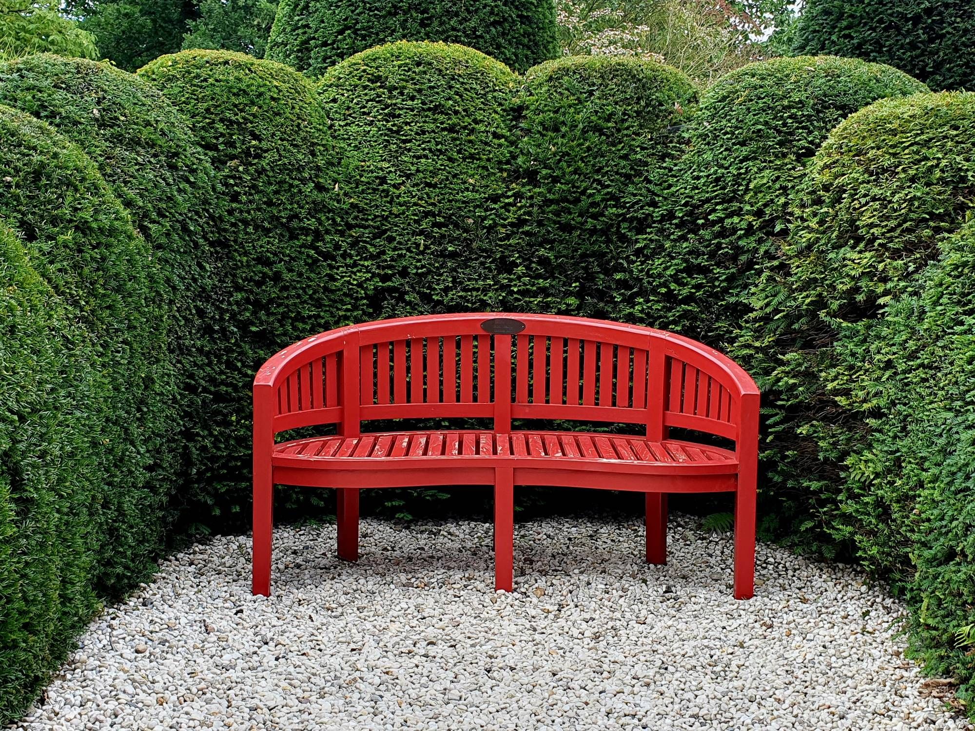 In einem Park: eine Sitznische mit einer leicht geschwungenen roten Holzbank. Ihre Form passt genau in die durch die Hecke gebildete Nische. Die immergrüne Hecke ist an der Oberkante in gleichmäßige Wellen geschnitten worden. Der Boden ist mit weißem Kies bedeckt.