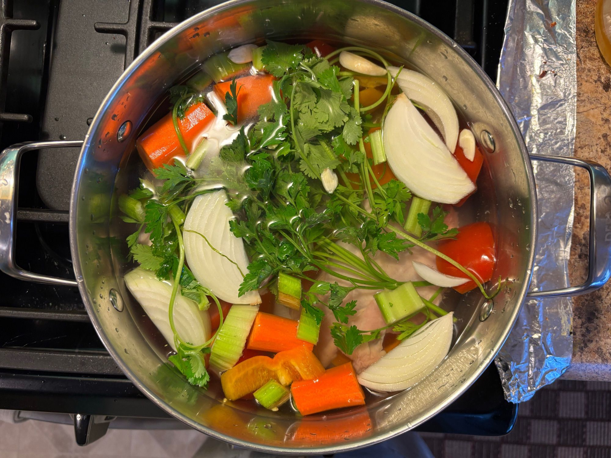 A cocotte filled with water, onions, carrots, garlic, celery, sweet peppers, tomatoes, and bone-in chicken, with stalks of coriander and parsley.