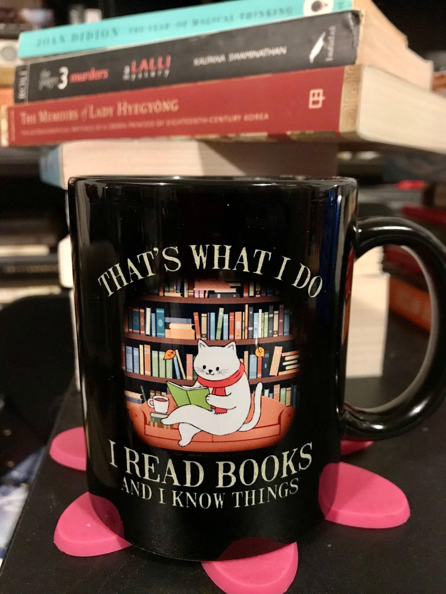 A black coffee mug with a cartoon of a smiling white cat, wearing a red scarf, engrossed in a green book, sitting on a peach chesterfield. Beside her, on the cushion are a couple of books, on which is a steaming cup of tea? coffee? snootled hot chocolate?.

The couch is in front of bookcases loaded with books. The words "That's what I do. I read books and I know things" are on the mug. The mug is on a fuchsia coaster, in front of a tower of books.