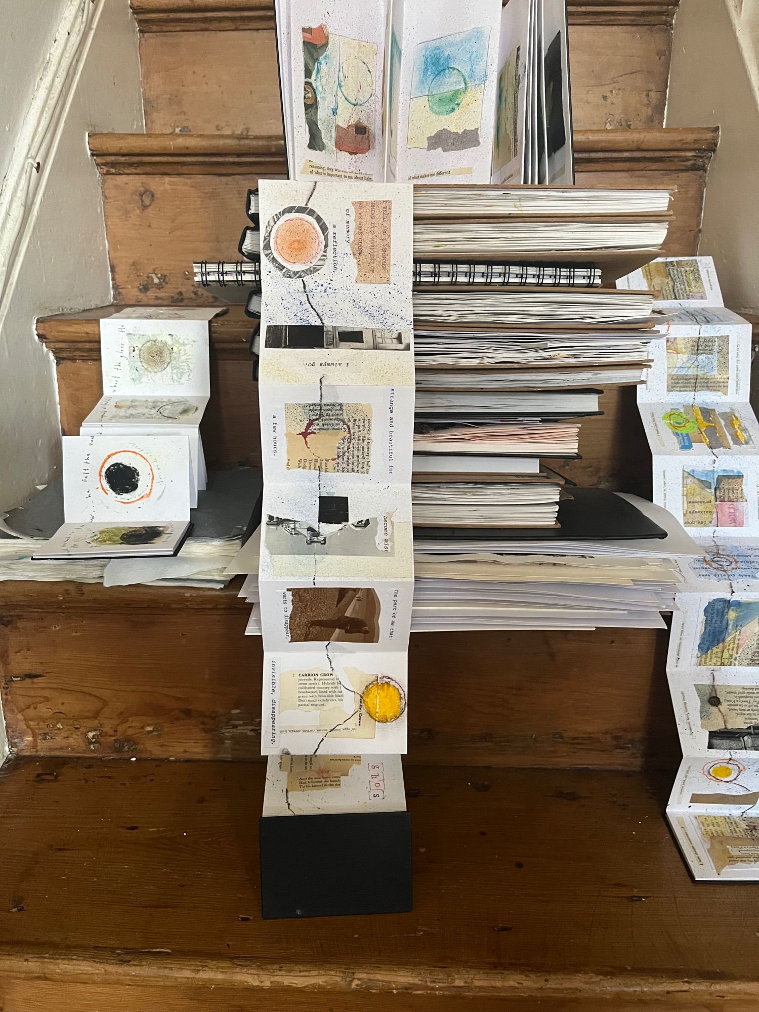 Image shows numerous notebooks arranged on staircase with concertinas notebooks cascading down the stairs. Showing collages of paint, paper scraps, bottle tops, wire, ink, fragments