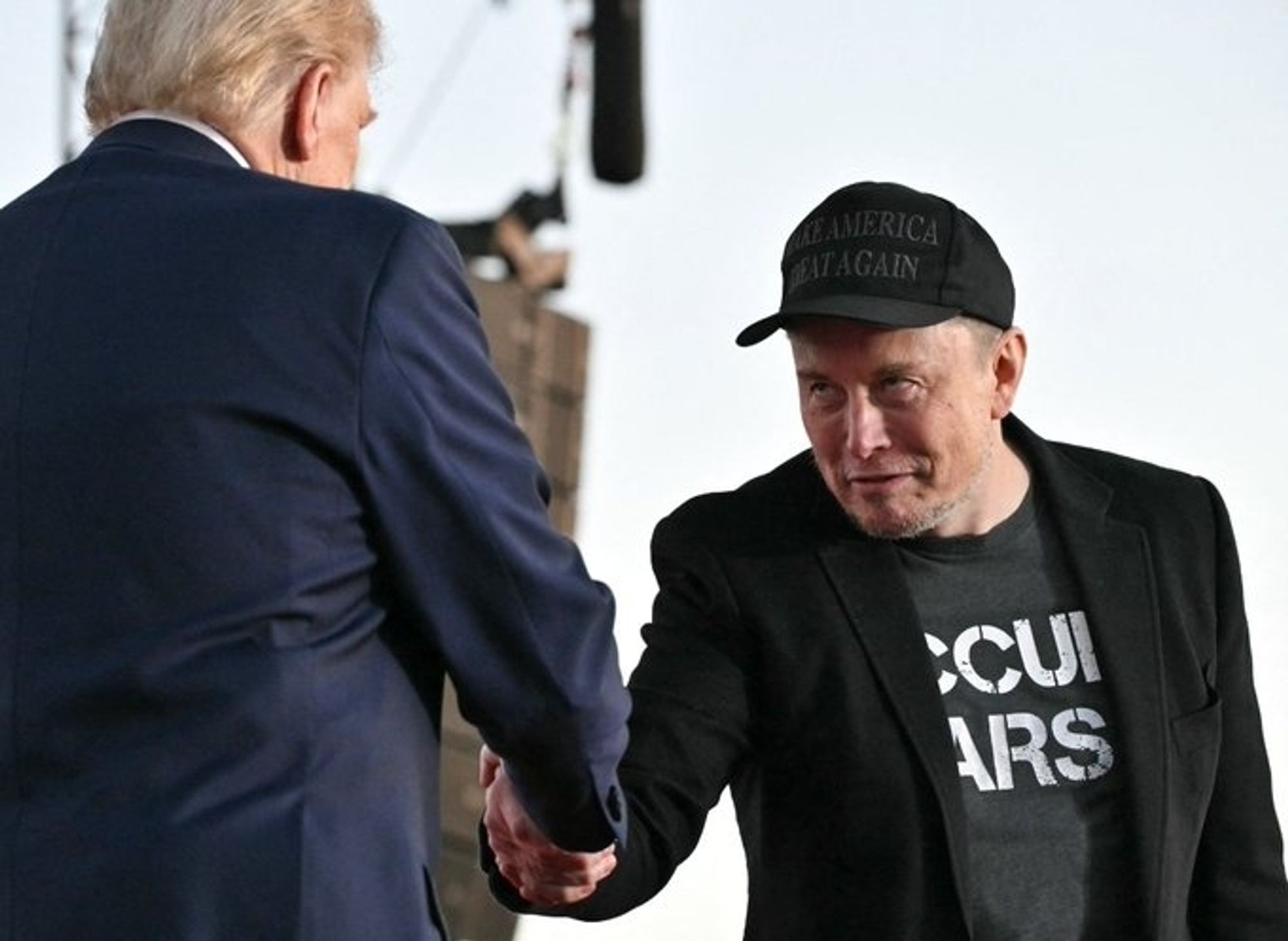 Elon Musk shaking the hand of Donald "Satan" Trump. He seems to be wearing a T-shirt bearing the slogan SCUM ARSE.