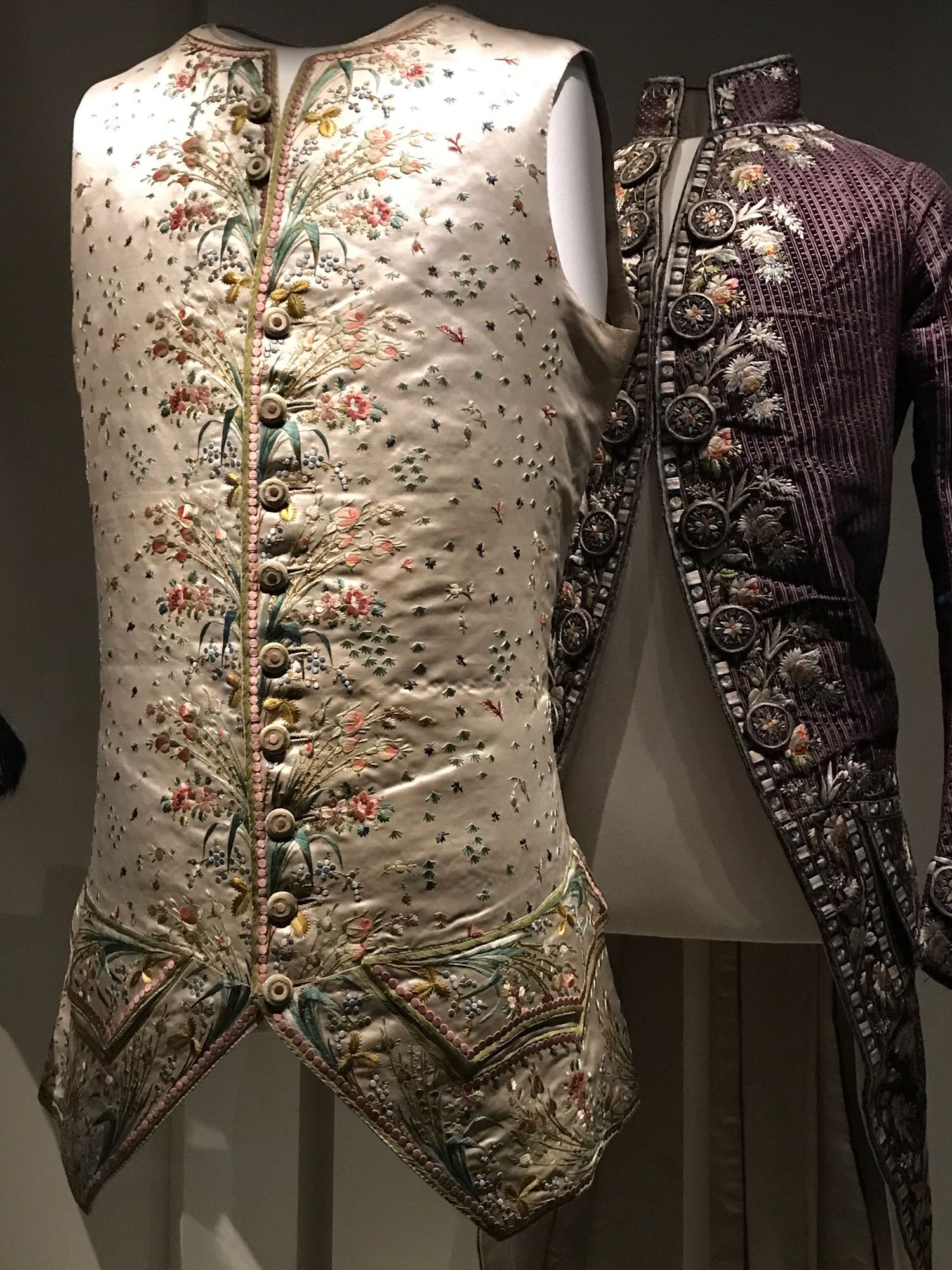 Champagne-computed waistcoat with fine floral embroidery in the front; in the background a lilac coat with large embroidered buttons and large floral embroidery