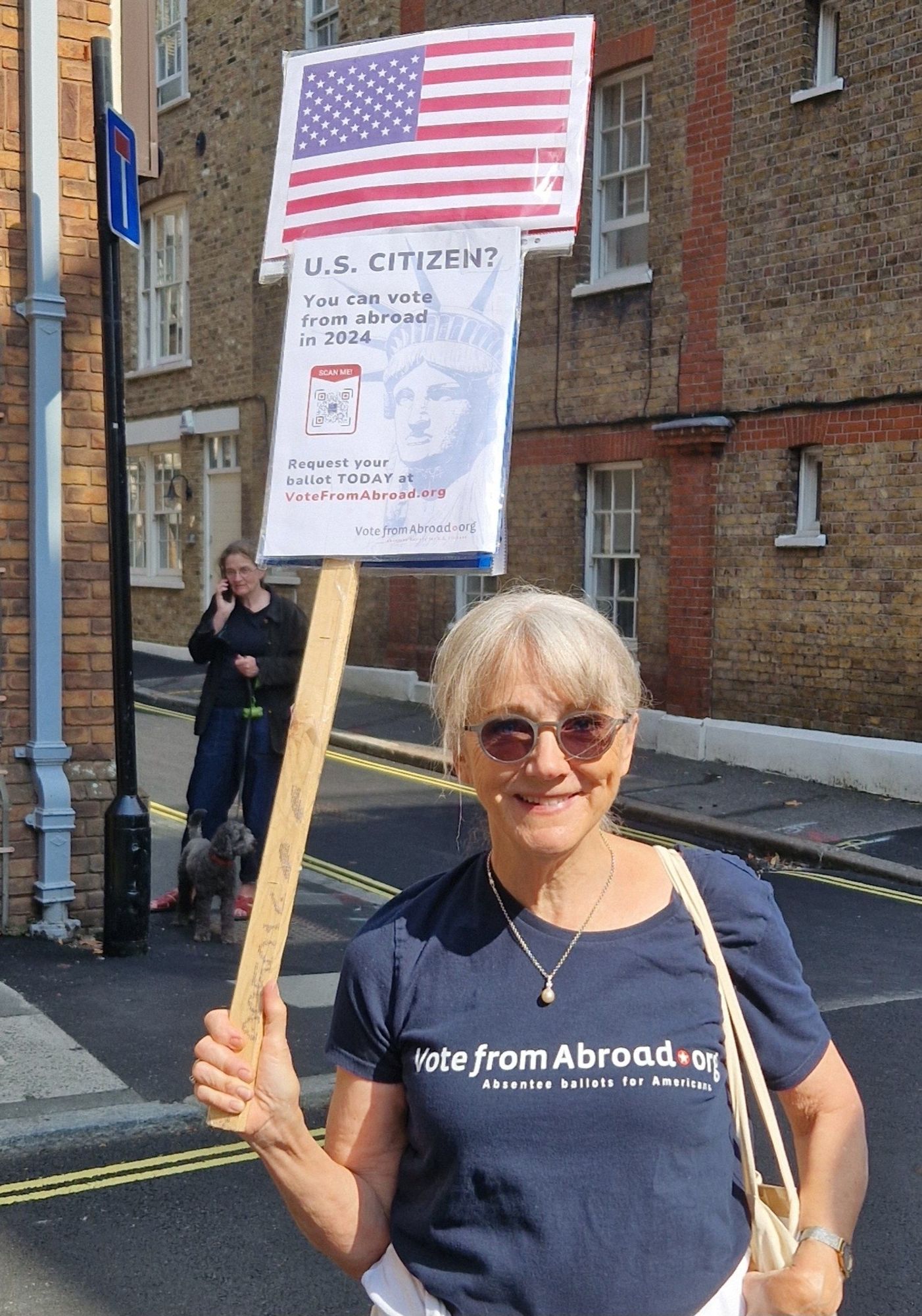 A beautiful smiling lady wearing a vote from abroad t-shirt, holding a sign with information about US citizens abriad being able to request their ballot.