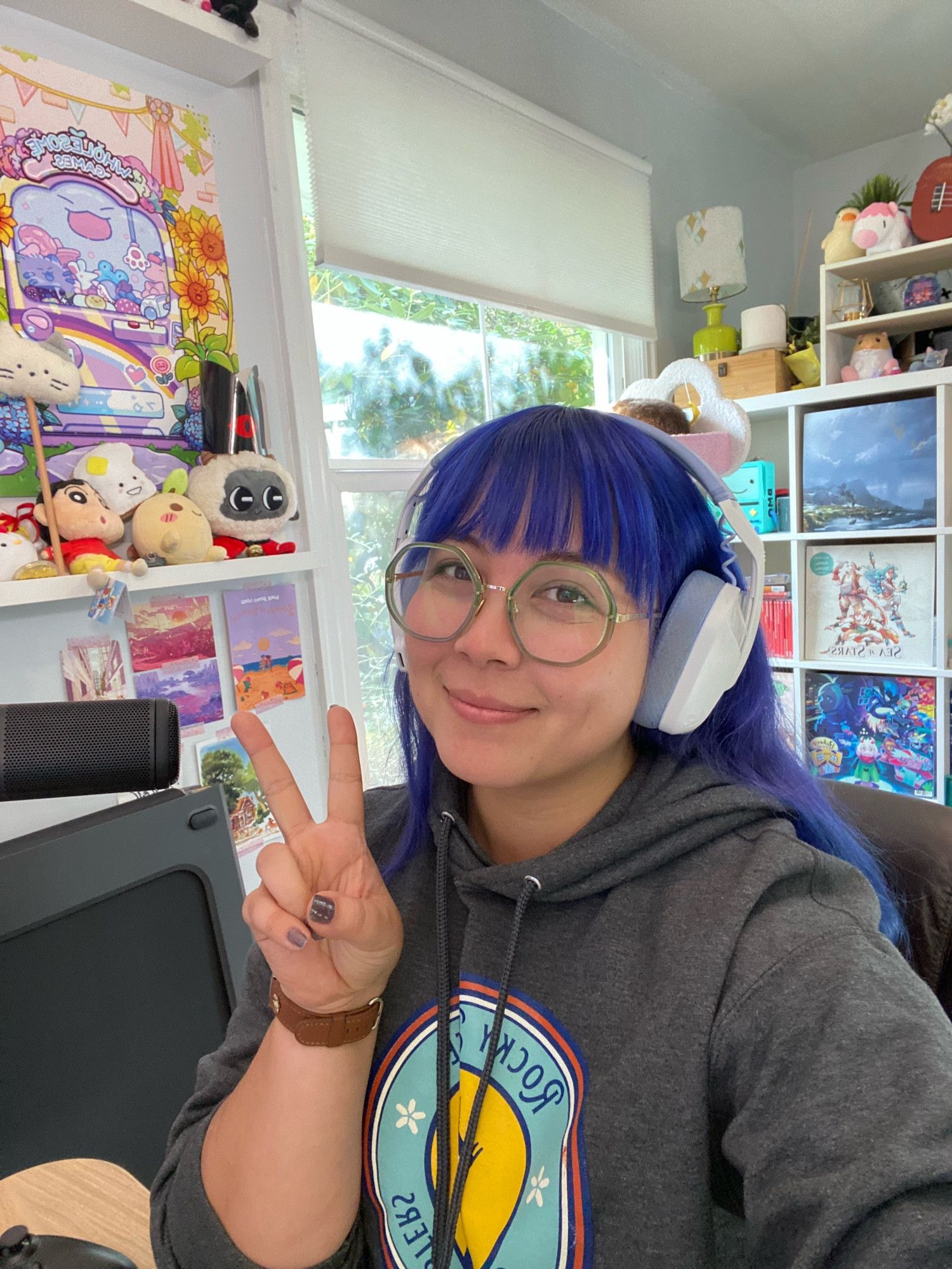 Jenny taking a selfie. She is in a room lit with natural light from an overcast day. Behind hee is a window. On the wall are postcards and art from various games, and there’s also shelving filled with game vinyls, plushies, and figures. 

Jenny is wearing a grey hoodie, green rimmed round glasses, and has purple-blue long hair with bangs. She’s also wearing white and lavender headphones. 