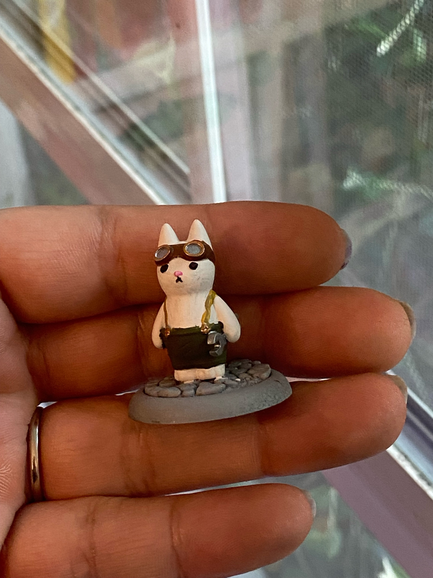 A photograph of a hand holding a 3D mini figure in front of a window. 

It is an (unfinished) painted cat figure holding a wrench and wearing suspender overalls and goggles standing on a cobblestone base. Jenny has painted the cat white, the suspenders mustard, the pants dark olive green, and the goggles brown, copper, and silver. The cobblestone is unpainted. 