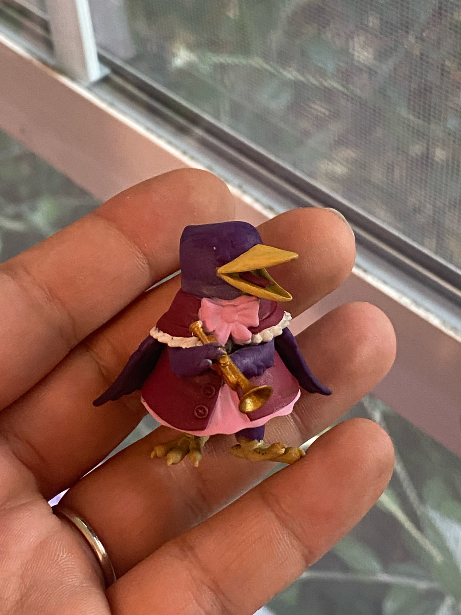 A photograph of a hand holding a 3D mini figure in front of a window. 

It is an (unfinished) Raven bard figure with no base. The raven is painted dark purple with a pink, red, and white cloak tied with a bow. It’s holding a trumpet painted gold.