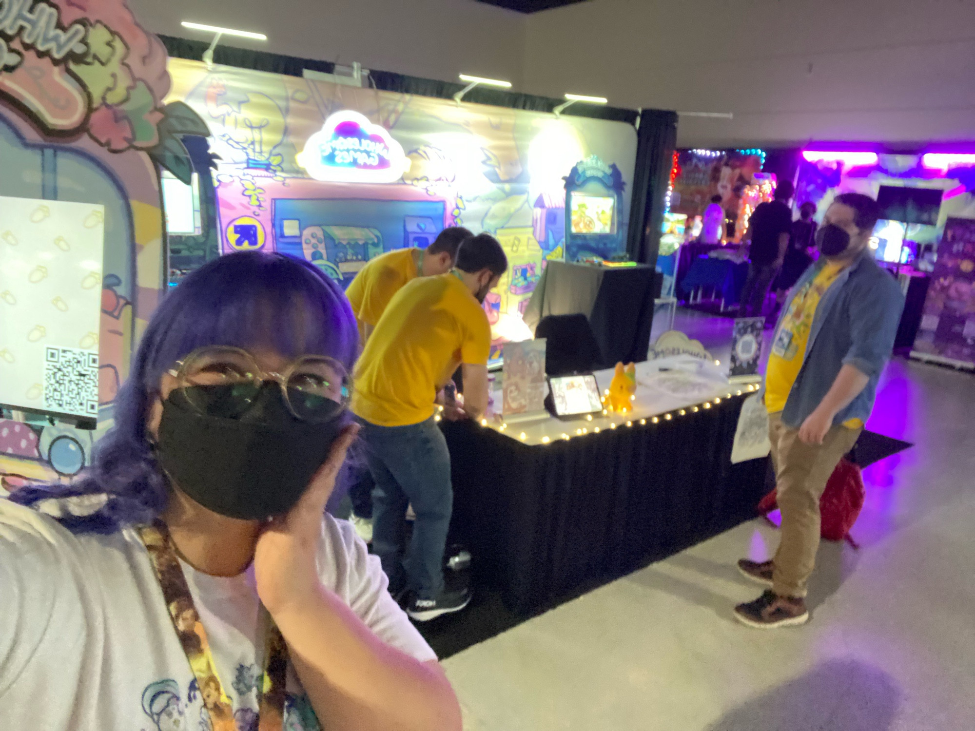 Jenny taking a selfie at the first ever physical Wholesome Games booth during set-up. 

In the background Evan and Matthew are taping lights to the table and James is looking over to see what Jenny is doing. 