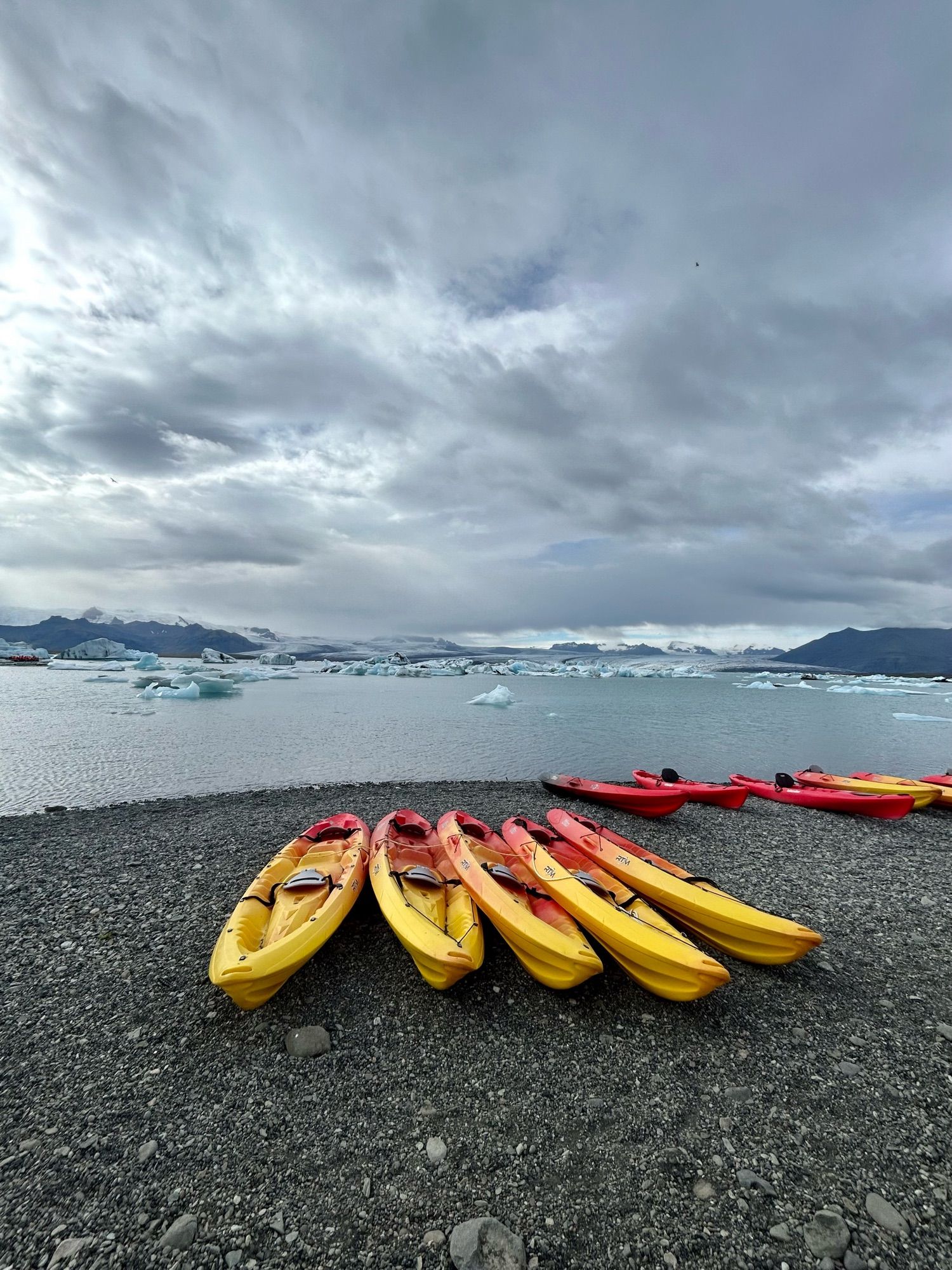 Jökulsárlón Glacier Lagoon in Iceland. Bright yellow and red sea kayaks are pulled up on a shingle beach under a gloomy sky. Small icebergs broken off the glacier in the background are floating in the middle distance