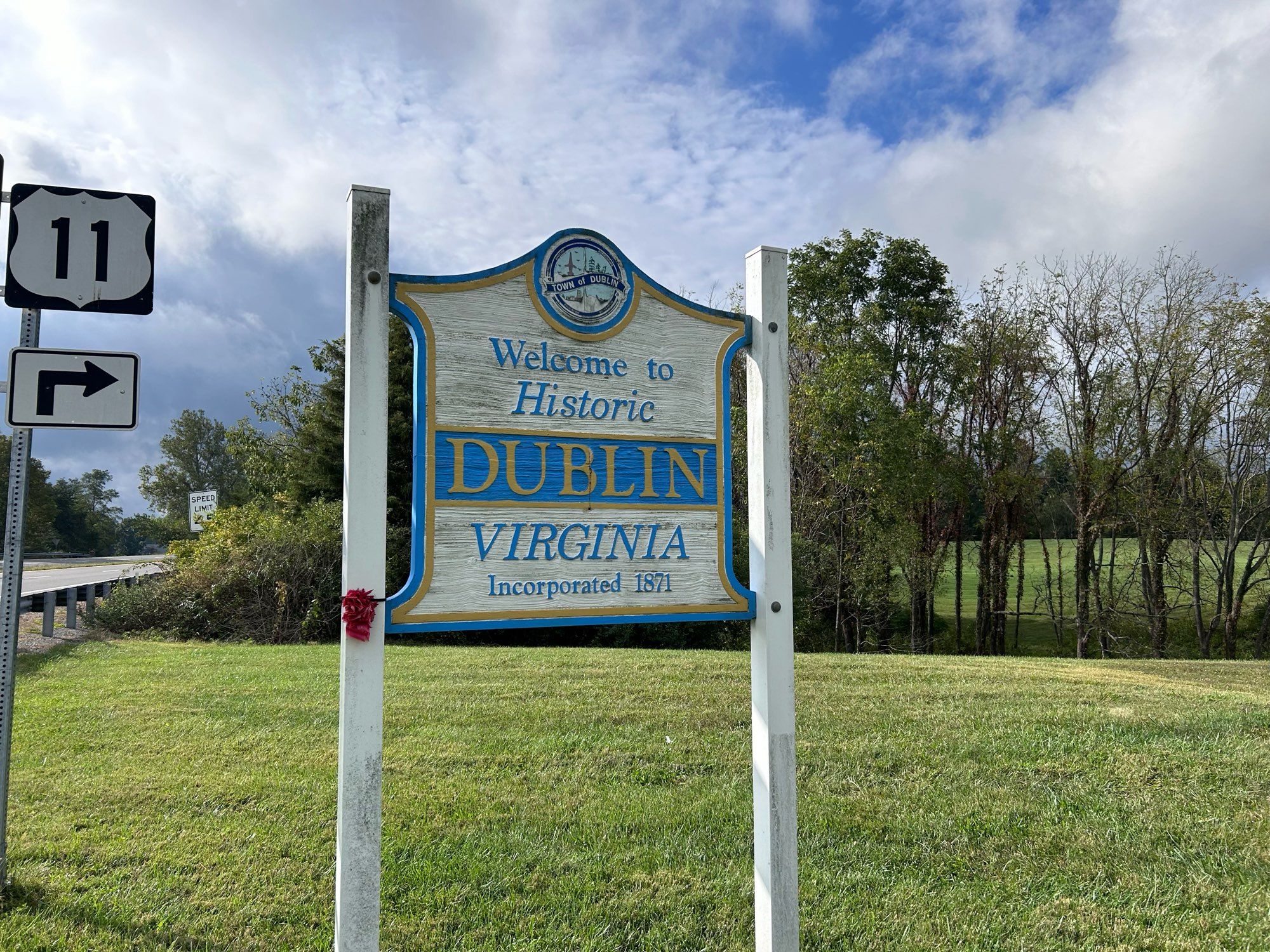 Roadside sign welcoming people to Dublin, Virginia. It is white and blue on a green field with a partly cloudy sky behind.