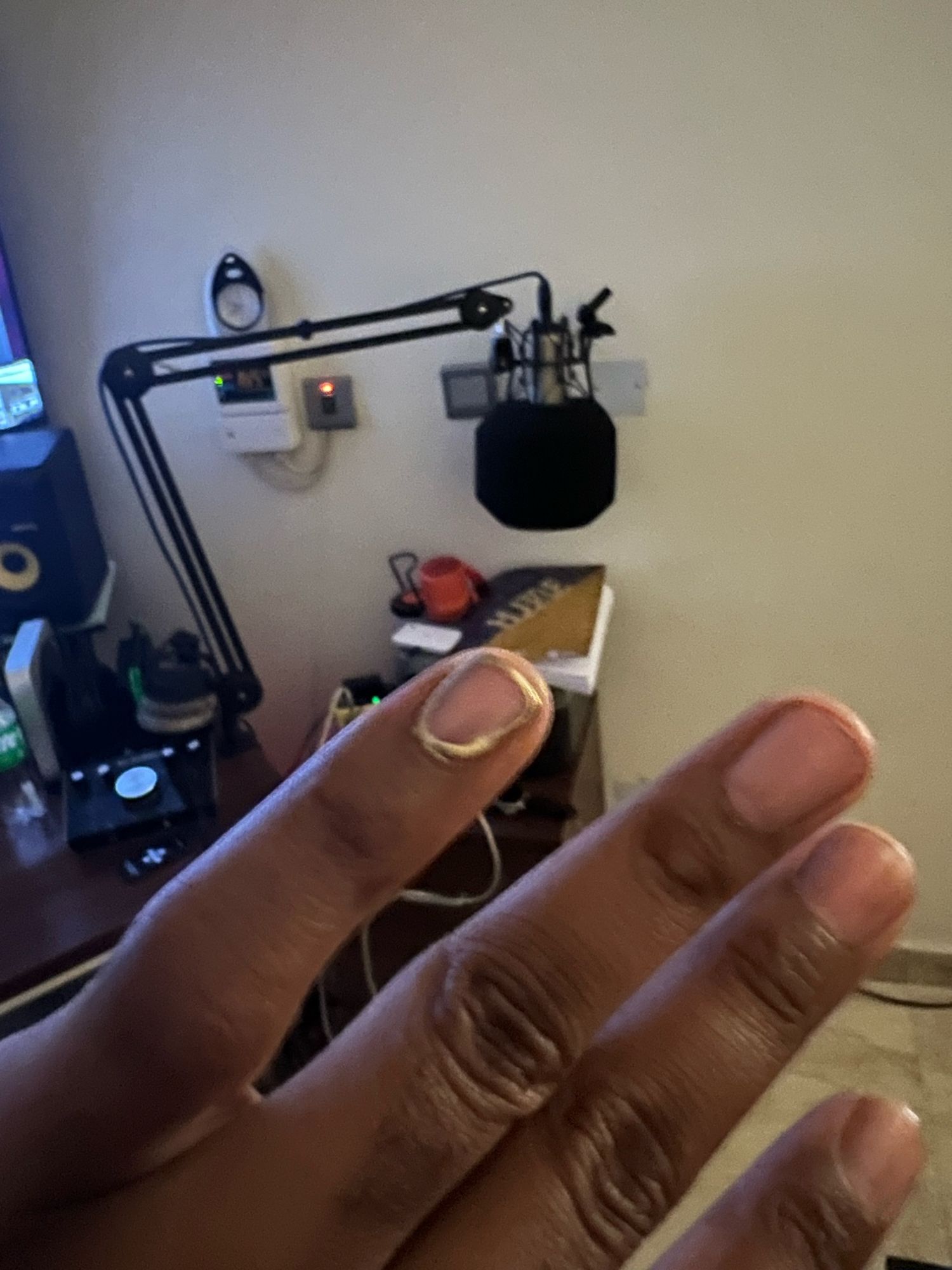 Picture of a black man’s left hand with the pinky nail of his crooked pinky painted around the edge with gold nail polish. The nail polish job is badly done. In the background, you can see studio equipment, and a microphone and a clock on the wall.