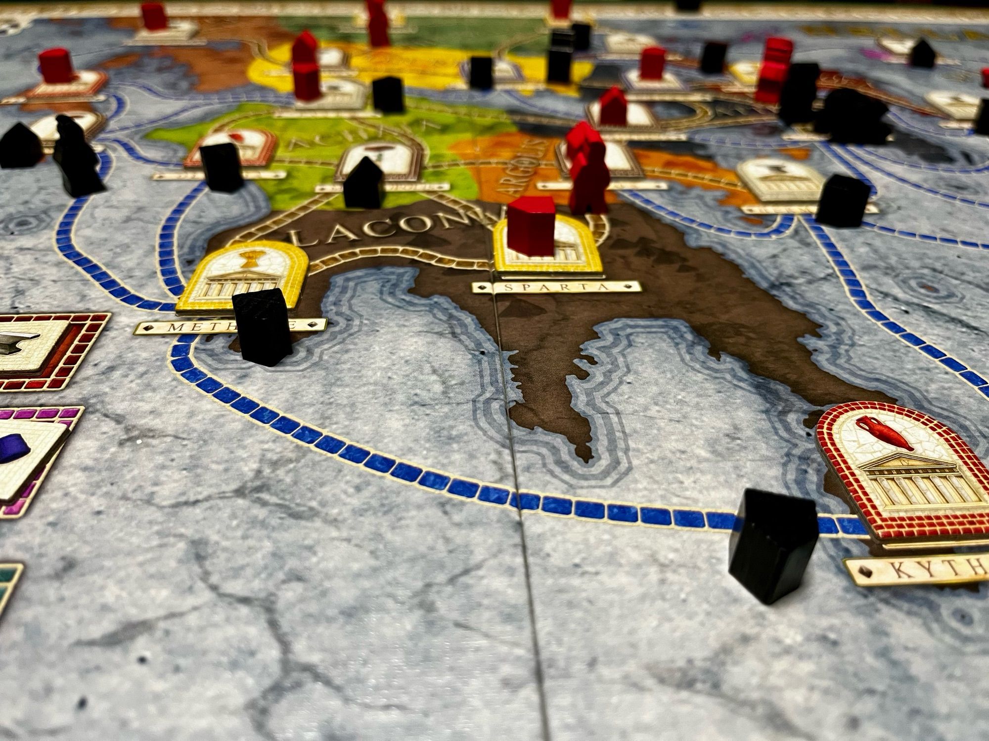 A close image of Concordia with the Hellas board, some blue and red wooden pieces and cardboard arch-shaped tokens representing production. Each region is linked via blue dotted routes for ships and brown for meeples.