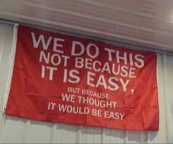 Red flag hanging on a white wall that says "we do this not because it is easy, but because we thought it would be easy"