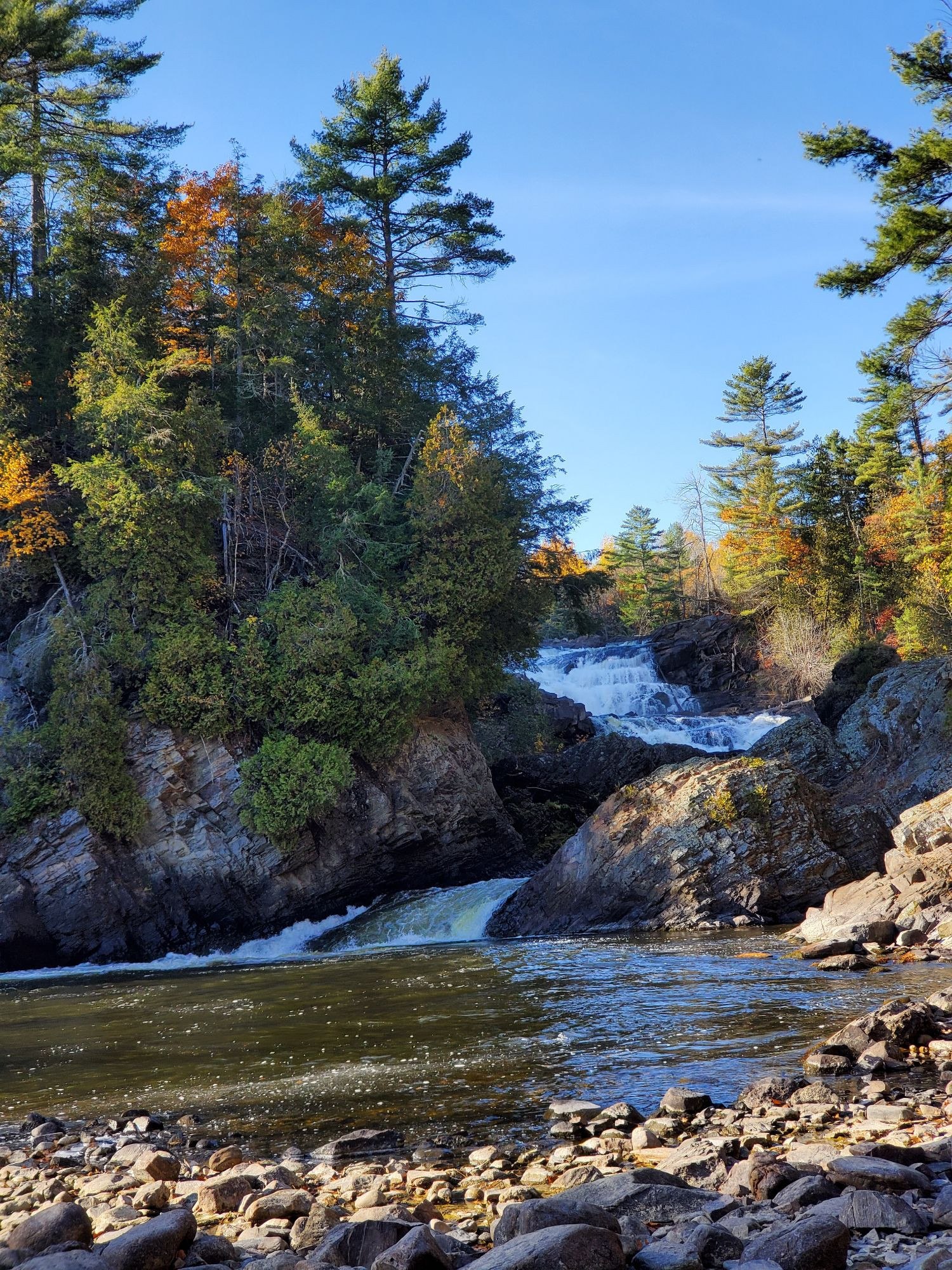 A waterfall as seen from shoreside downriver with trees on large boulders each side. 