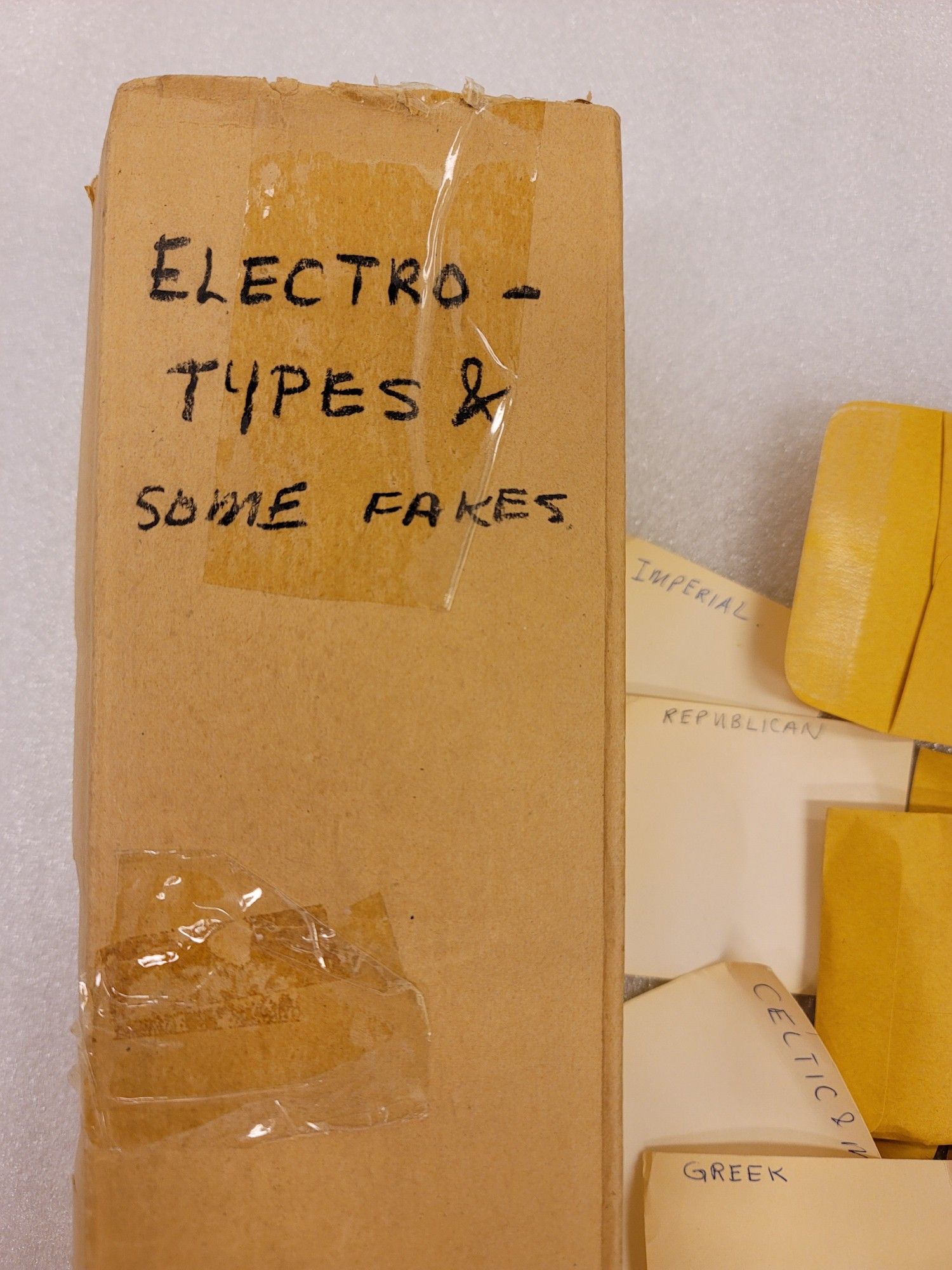 Brown cardboard box labelled "Electrotypes & some fakes." To the right a small pile of manila envelopes and browning bits of paper with Greek, Celtic, Republican, and Imperial, as labels.