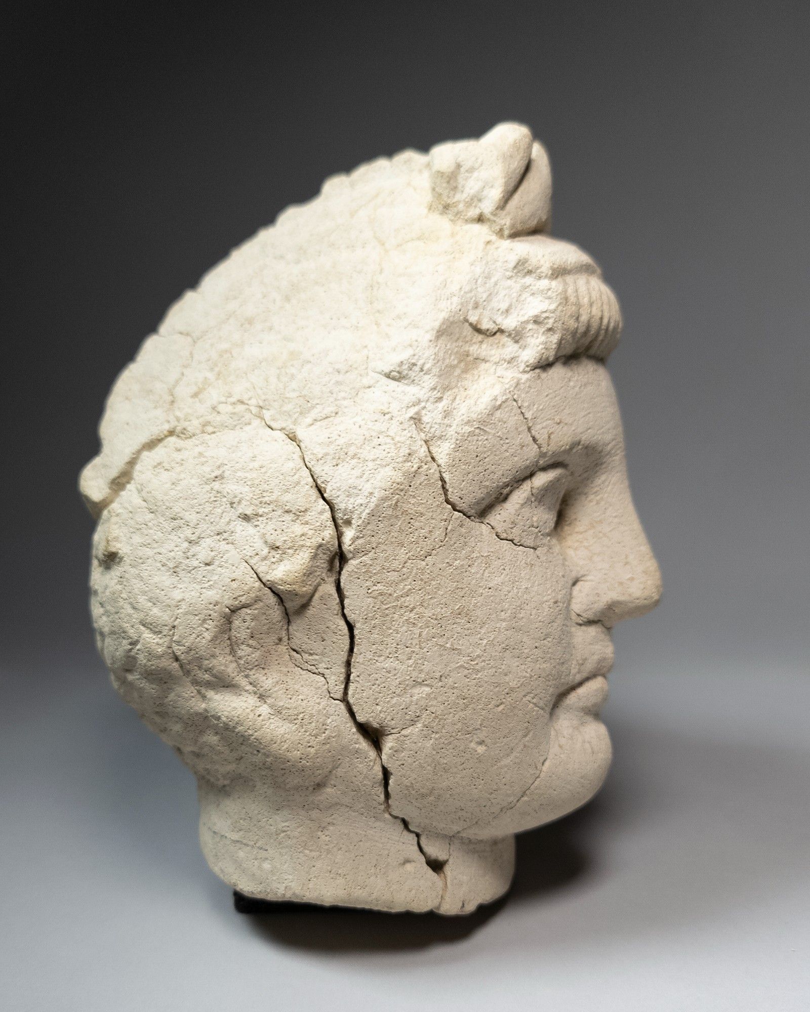 Profile of a light beige limestone head of a man facing to the right, on a grey background. Many cracks radiate from eye and side of the face near the ear. ROM958.61.87