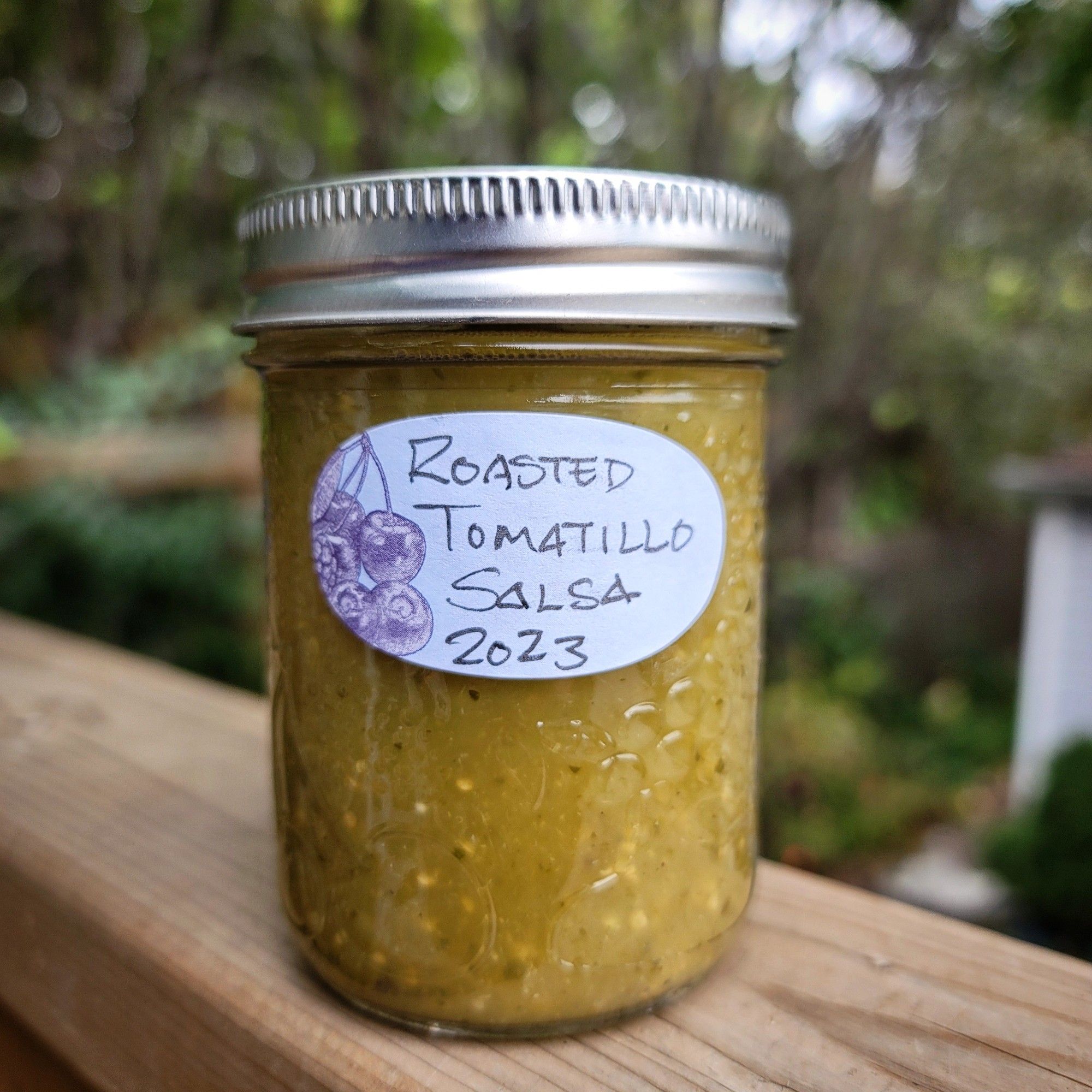 One glass canning jar of roasted tomatillo salsa on a wooden deck railing overlooking the blurred backgroung garden.