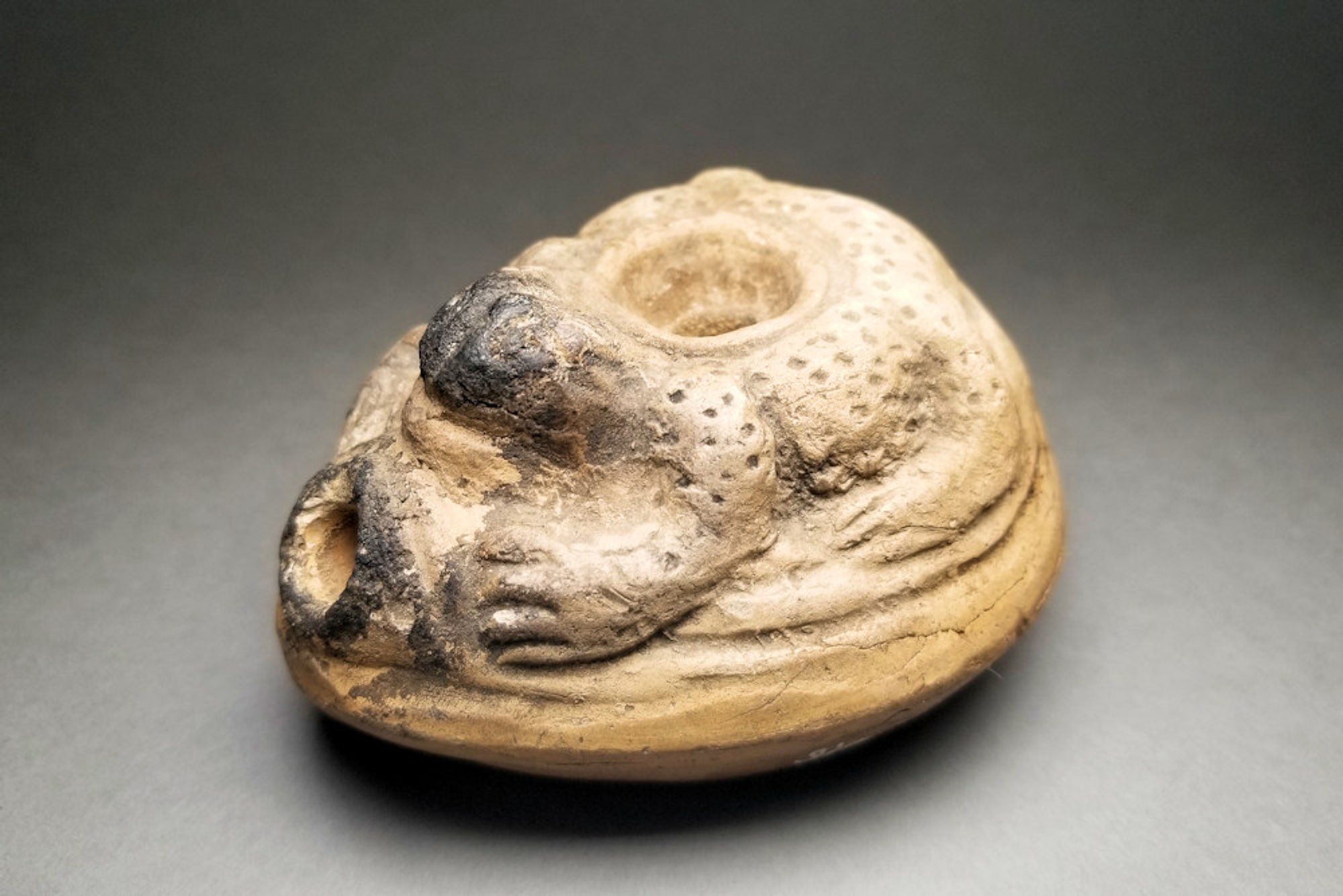 Roughly almond-shaped pottery oil lamp, buff-beige colour, with a frog on the top, its arms holding each side of the nozzle where a lit wick would have burned. Black, sooty deposit surrounds the nozzle and all over the face and head of the frog located just above the nozzle.