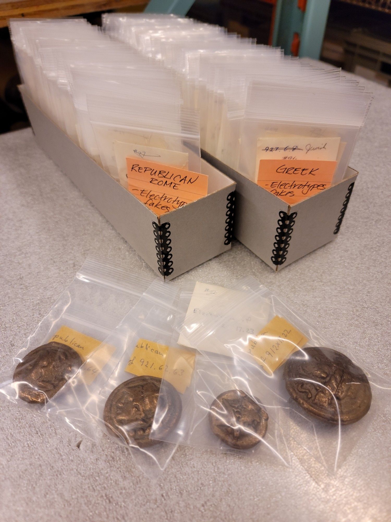 Four eletrotype Roman Republican bronze coin replicas in polyethylene bags arrayed in front of two grey archival boxes containing many more specimens.