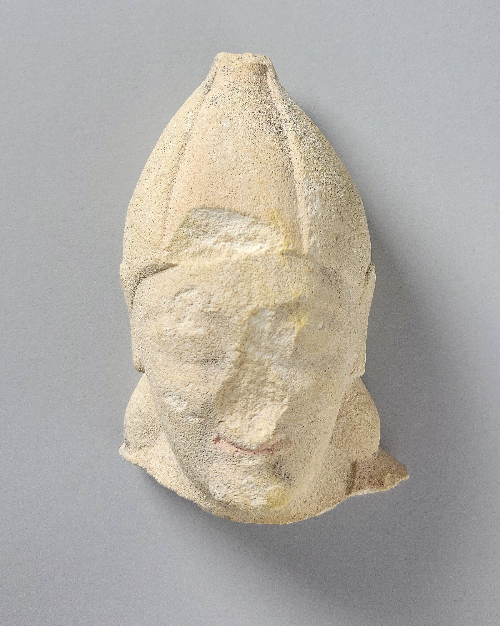 A small, 11.3 × 7.3 × 5.4 cm, head from a human figure. Roughly oval, wearing a close-fitting, conical, hat. Face is chipped and battered on chin, cheeks, and is missing its nose. Oval eyes. The mouth, with closed lips, is smiling, in a typical "Archaic smile."