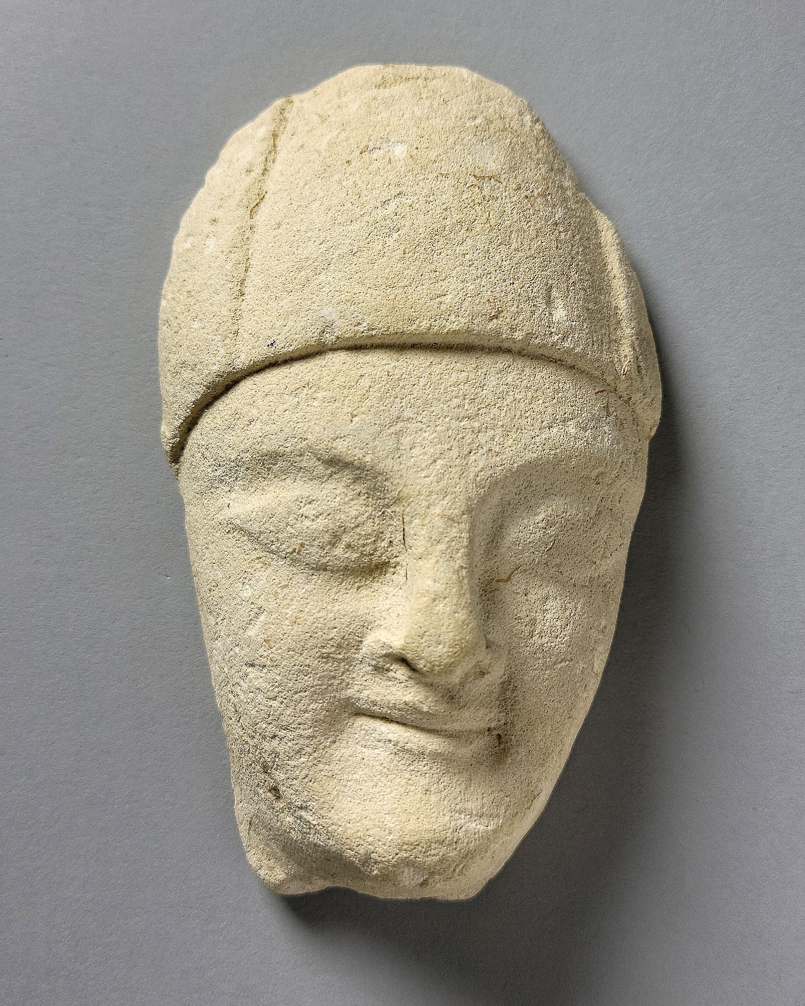 Oval-shaped head fragment of a limestone figure. The person wears a close-fitting hat, has almond-shaped eyes and their mouth, with closed lips, turns up subtly at the corners in a typical "Archaic Smile."