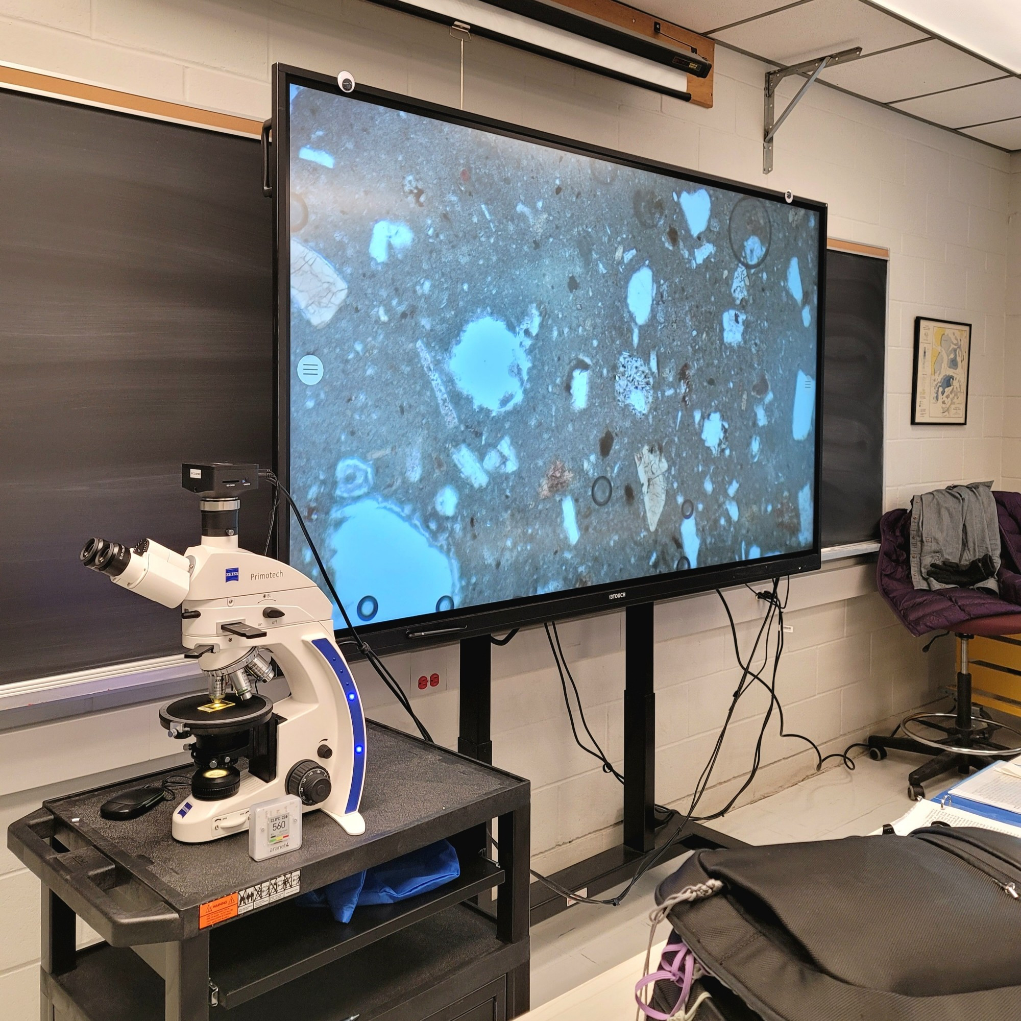 Big screen monitor attached to a microscope showing a ceramic thin-section petrofabric at 100X magnification, various rocks and minerals scattered around the screen.