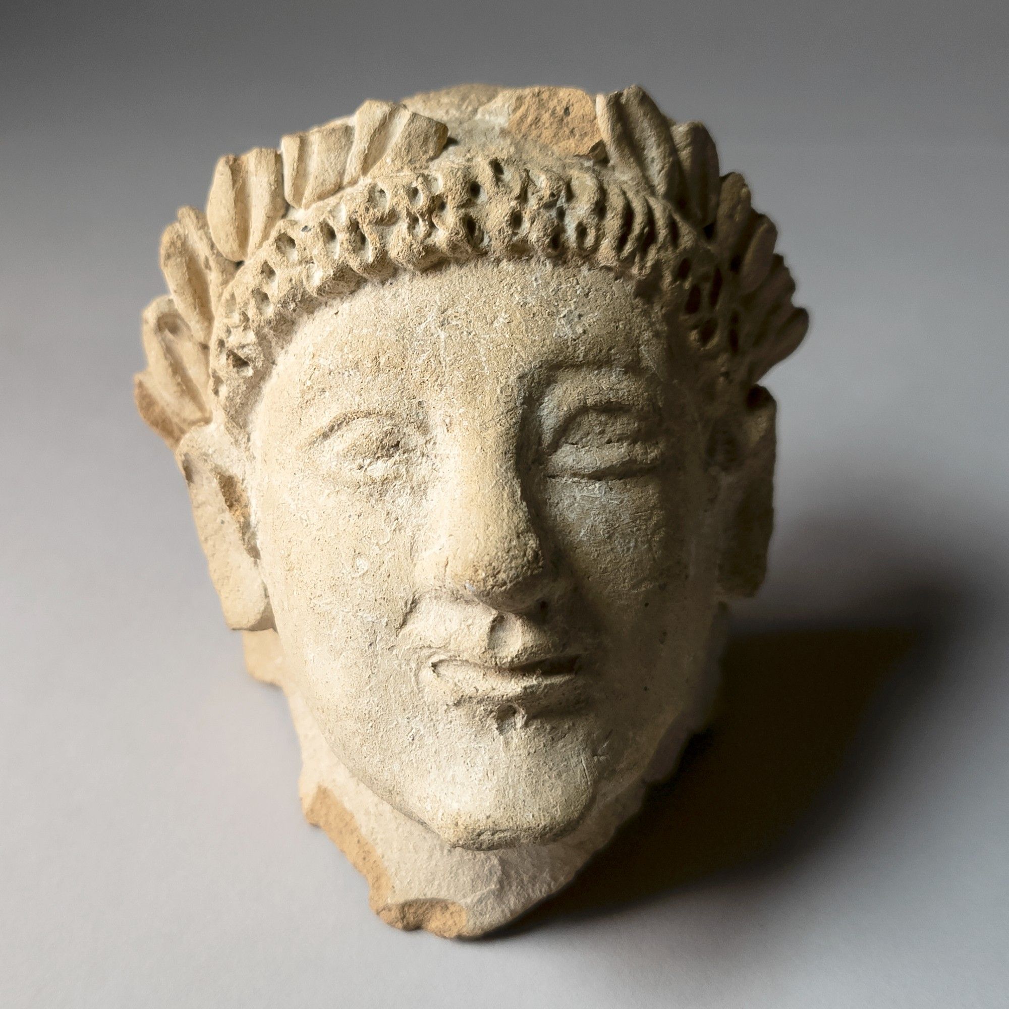 Head of a fragmentary ceramic figure, facing frontal, wearing a laurel wreath, lips slightly pursed in an "Archaic Smile," small indents or divots, in the middle, above upper lip and below lower lip. Grey background. ROM958.61.126