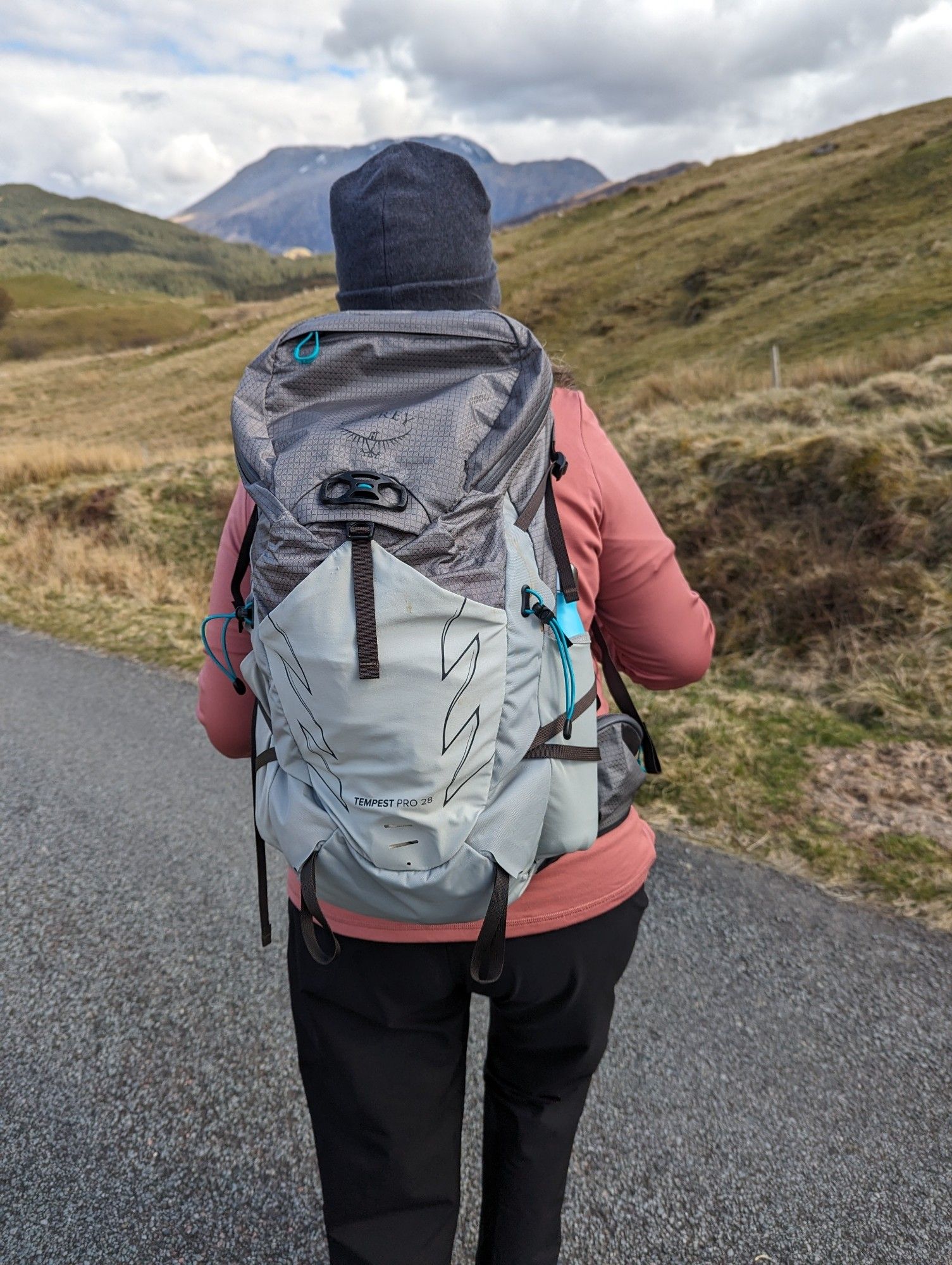 A middle aged woman with her back to the camera, in black walking trousers, soft pink top, dark grey hat and carrying a backpack is walking towards hills