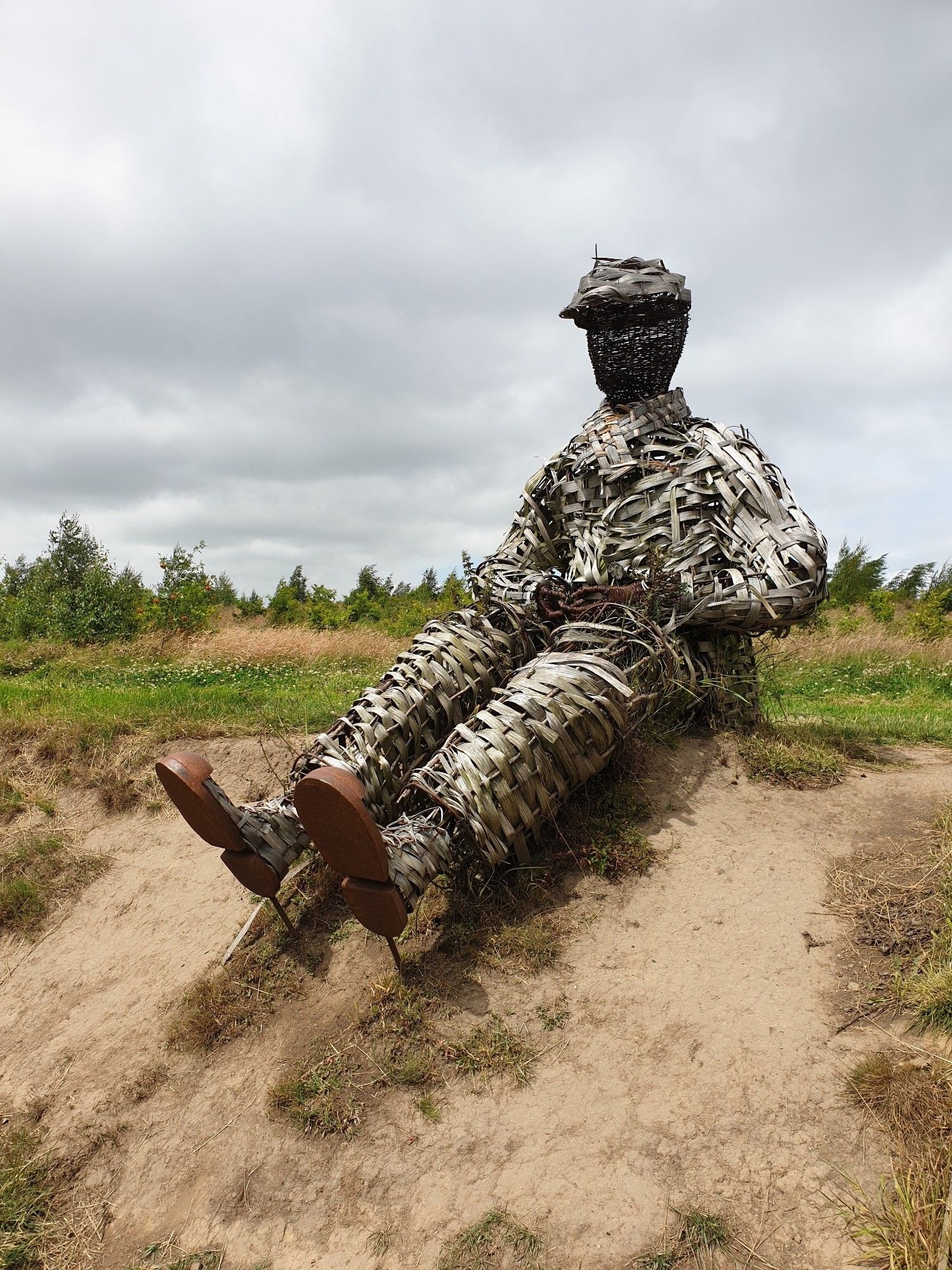 A wicker man seated on grass with his legs hanging down a sandy bank