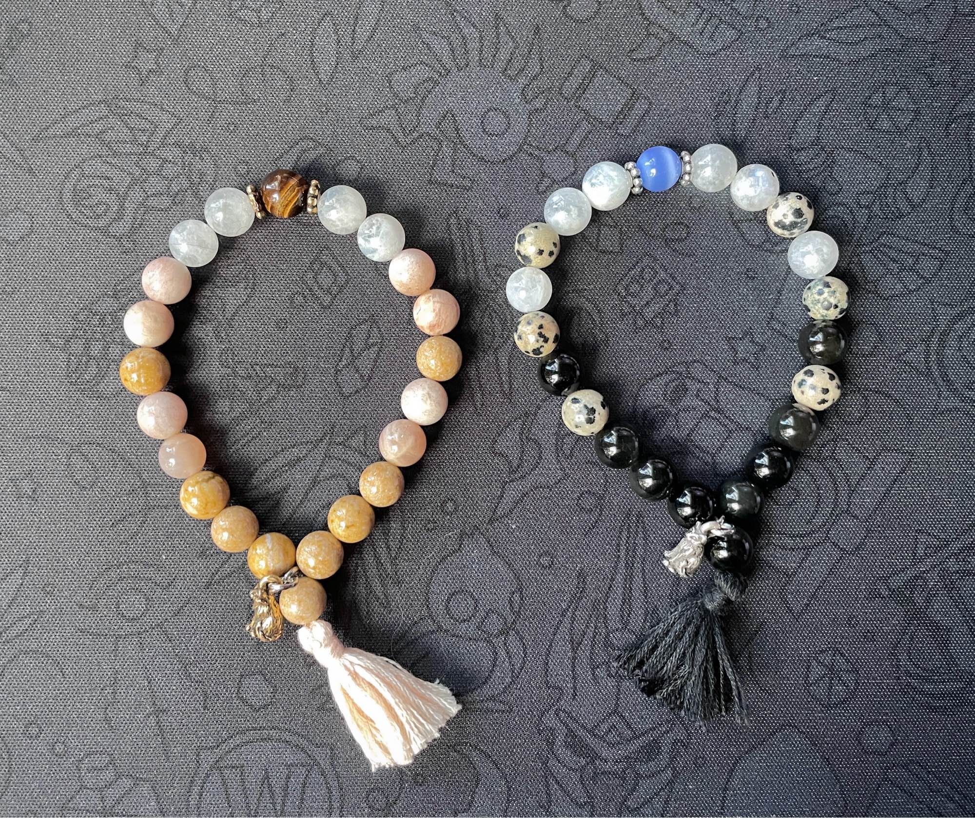 A cream colored and a black and white speckled bracelet made from 8mm beads, each with a tassel and small cat charm.