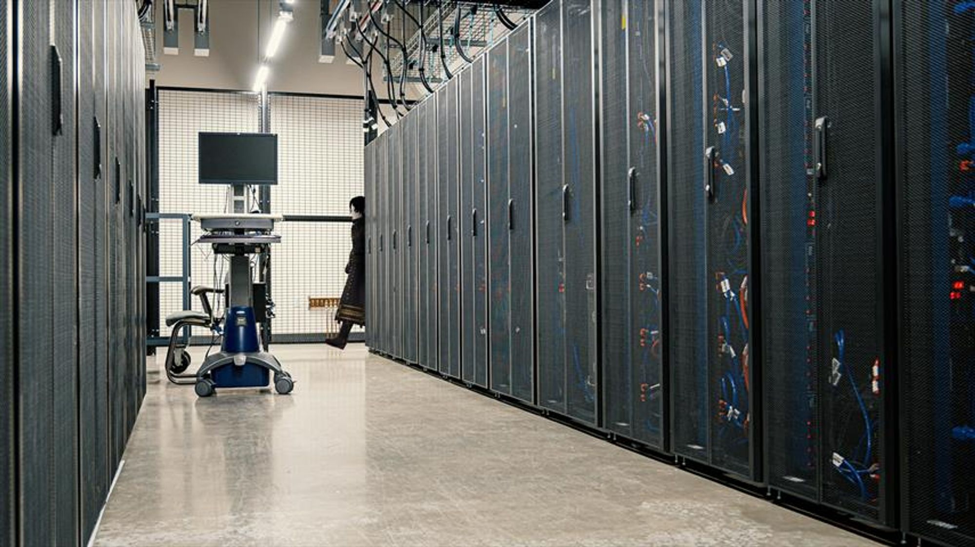 A person, partially visible behind a server rack, appears to be walking past or towards an aisle in a data center. They are wearing dark clothing and shoes.