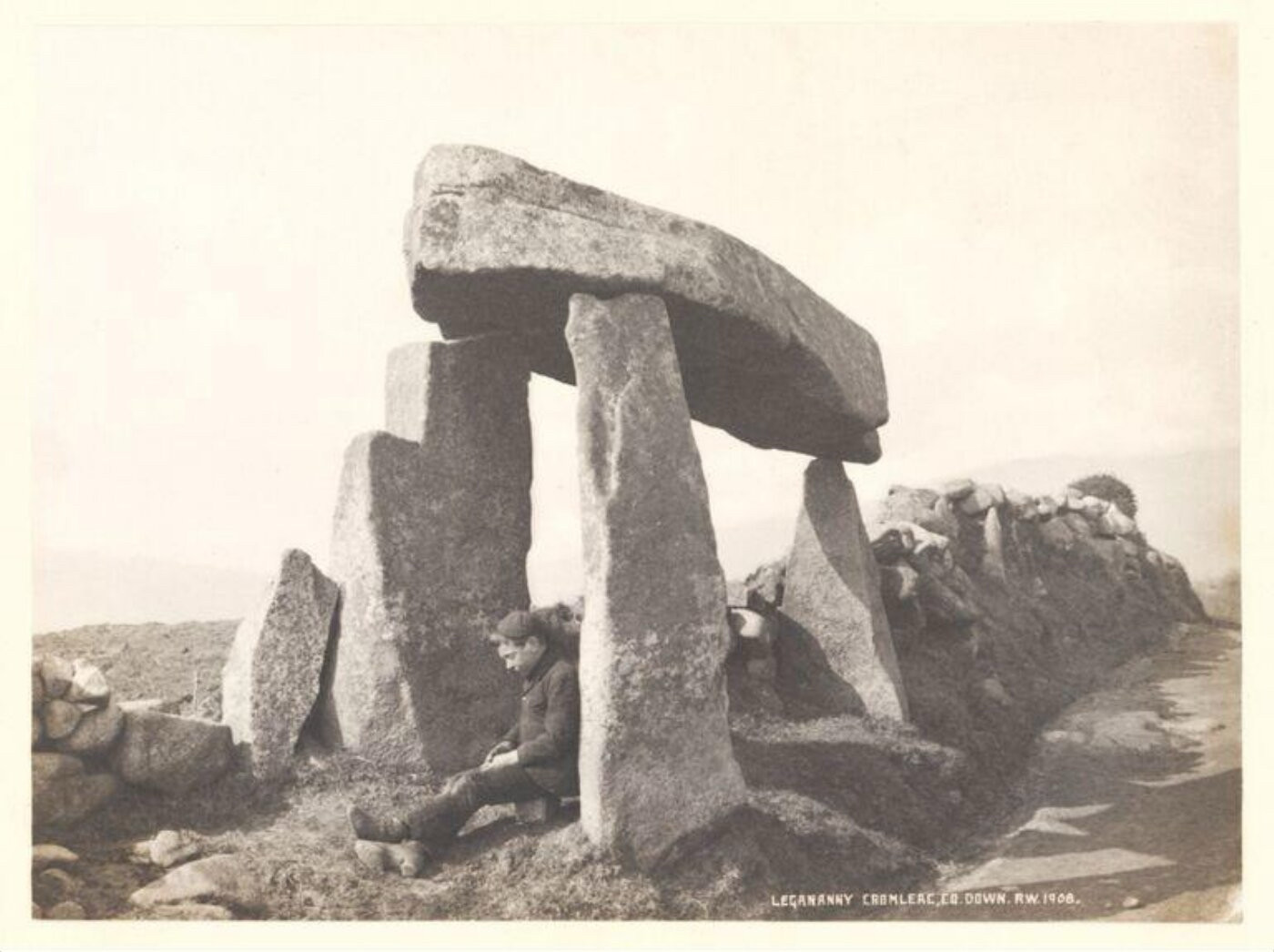 b&w print of a boy seated under a tripod arrangement of standing stones