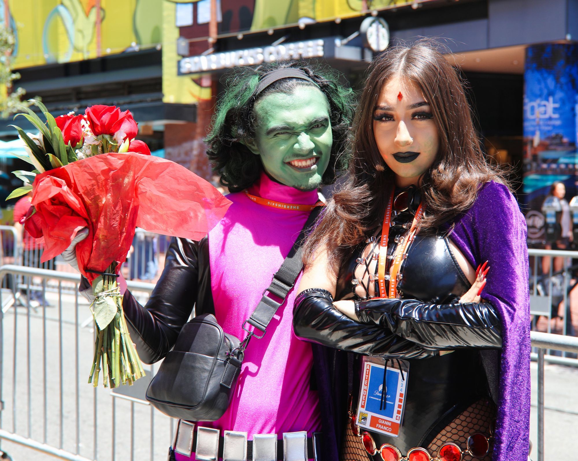 Cosplayers portraying Beast Boy and Raven in the middle of the street.  Young man in green face paint and pink and black tighs with white belt.  Young woman in leather arm bands and purple cape with arms crossed over cleavage.