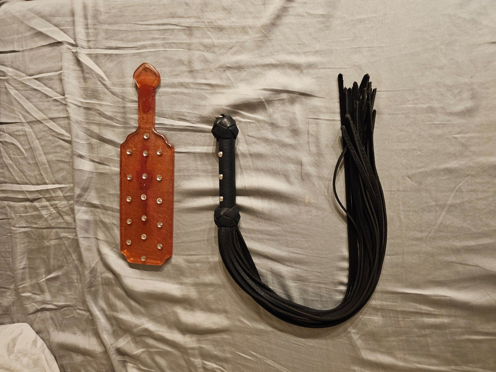 A pink resin paddle with thumbtacks and a black leather flogger