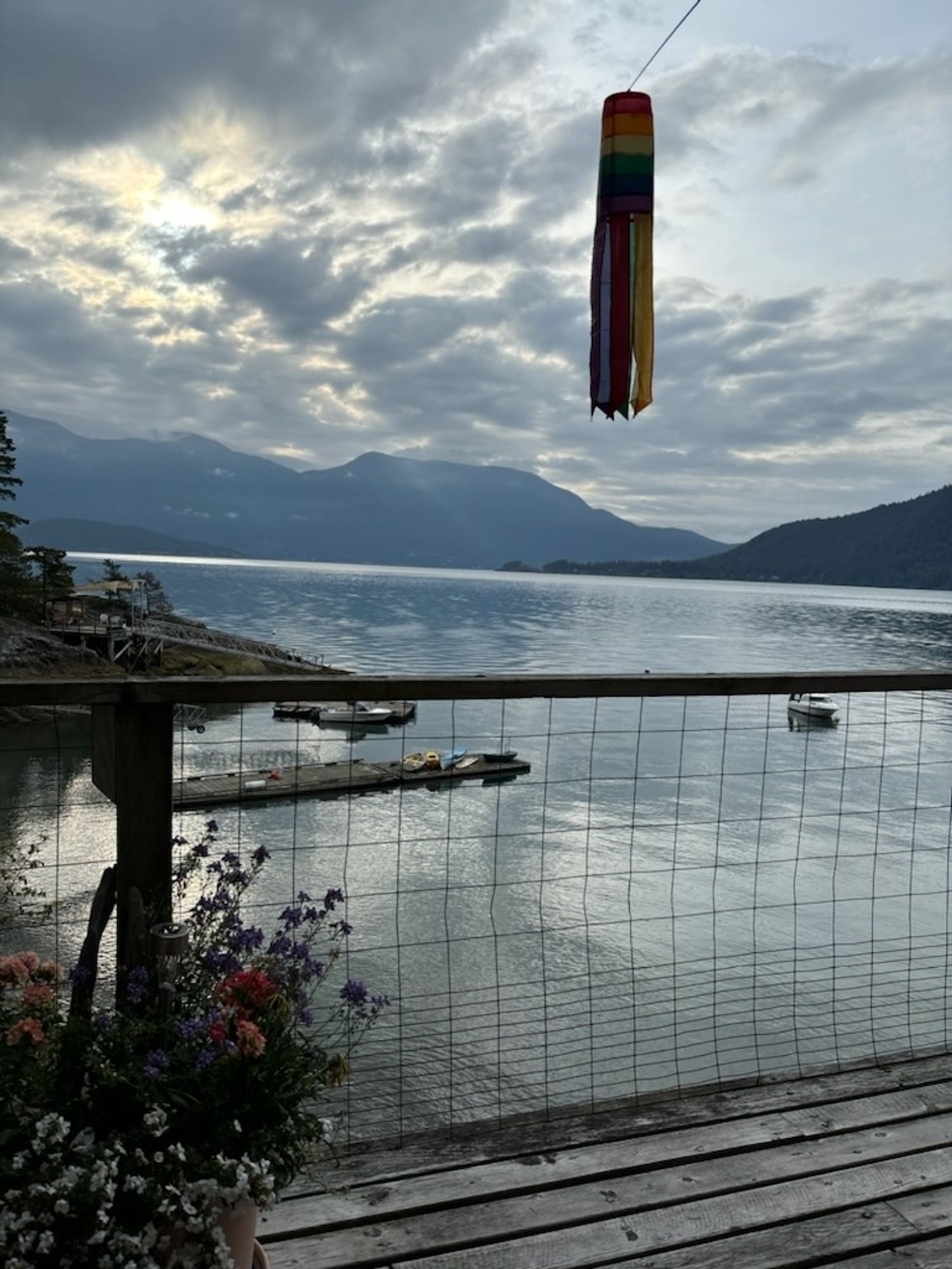 Cloudy sky above mountains and ocean. Above a wooden deck, there is a rainbow wind catcher. On the deck to the left is a planter of flowers