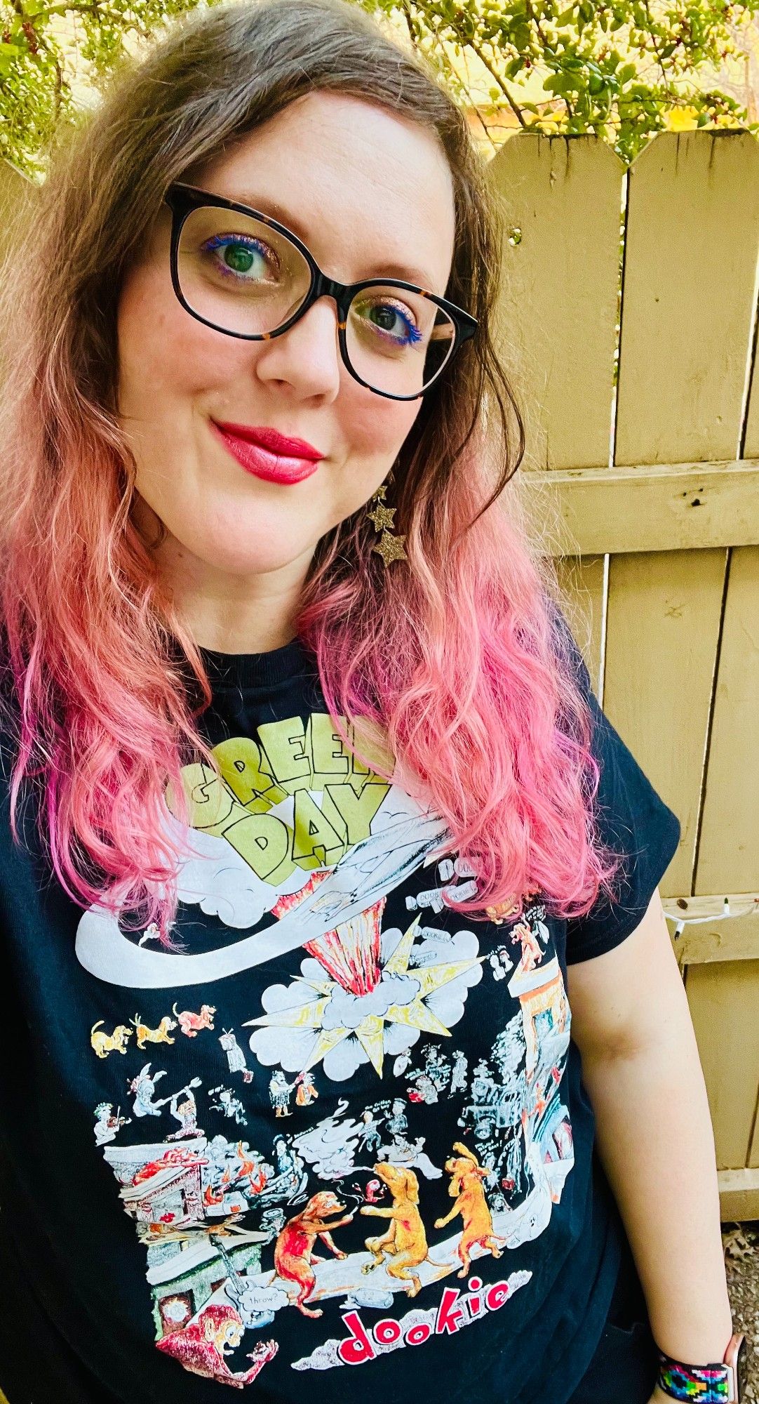 Selfie of me smiling with my mouth closed. I have brown and pink hair. I am wearing black and gold glasses, a Black Green Day Dookie album cover t-shirt, and gold star earrings.