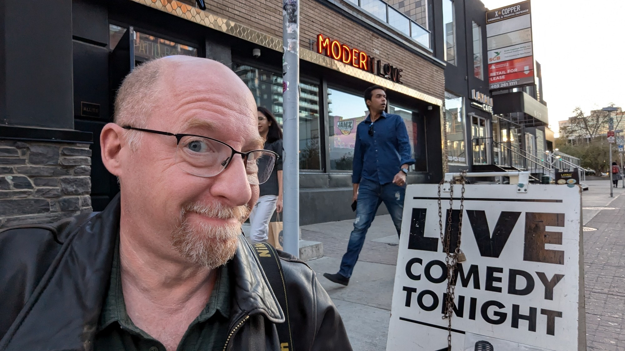 Bald Scott with glasses in front of a "Live Comedy Tonight" sign in front of a Calgary bar.