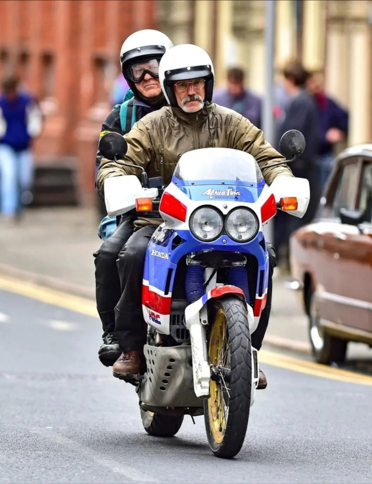 Sean Bean and Daniel Day Lewis on a bike both are wearing helmets. DDL has a moustache and glasses