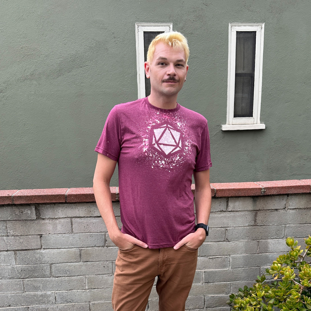 Xander is a bleach-blonde man with a dark mustache looking directly at the camera. His hands are in his pockets, and he's wearing a maroon tee shirt with a D20 design created with bleach.