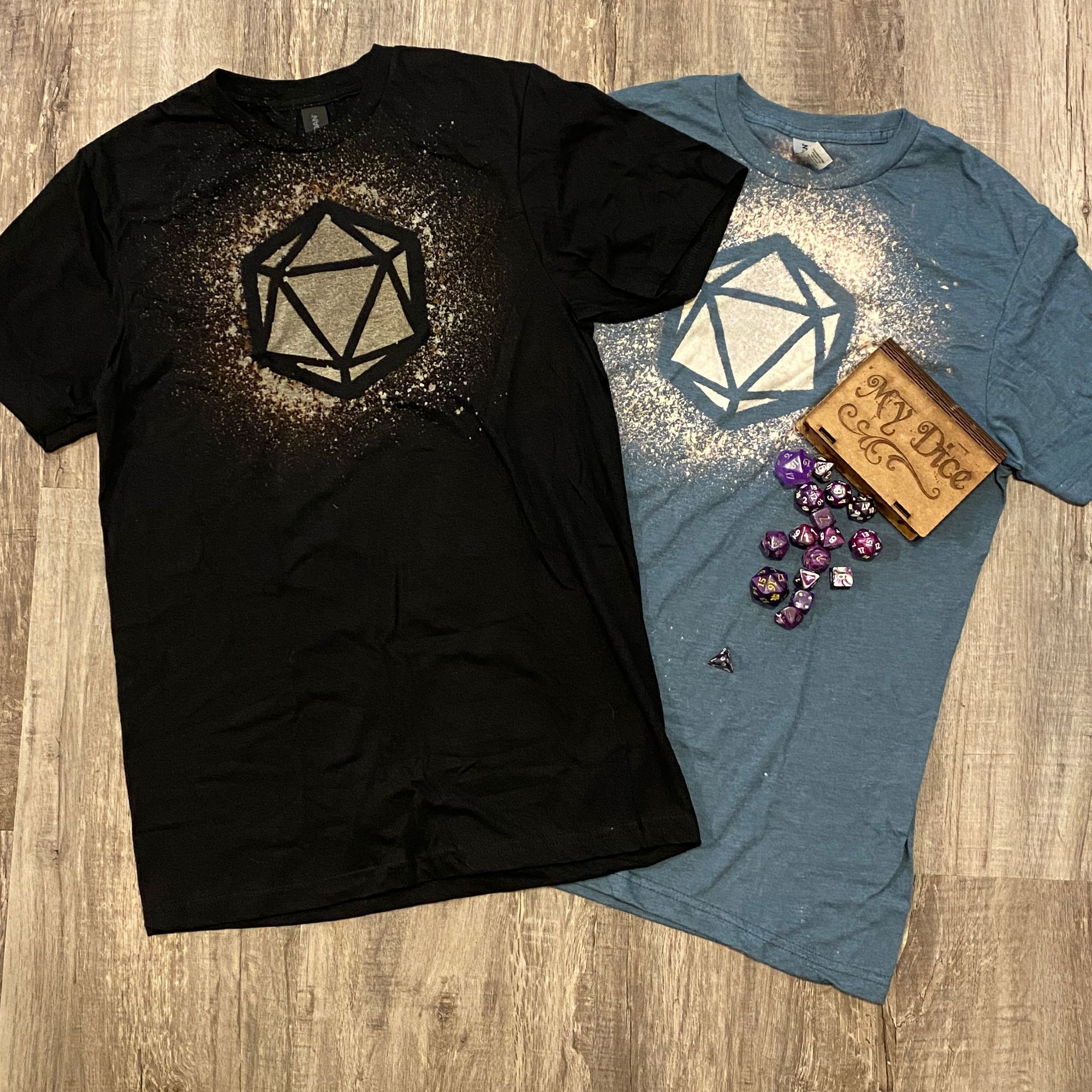 A black tee shirt and steel blue tee shirt lying on a light wood floor. Both tee shirts have a D20 design created with bleach. There is a wooden dice box that says "my dice" on the blue shirt with lots of purple dice spilling out of it.