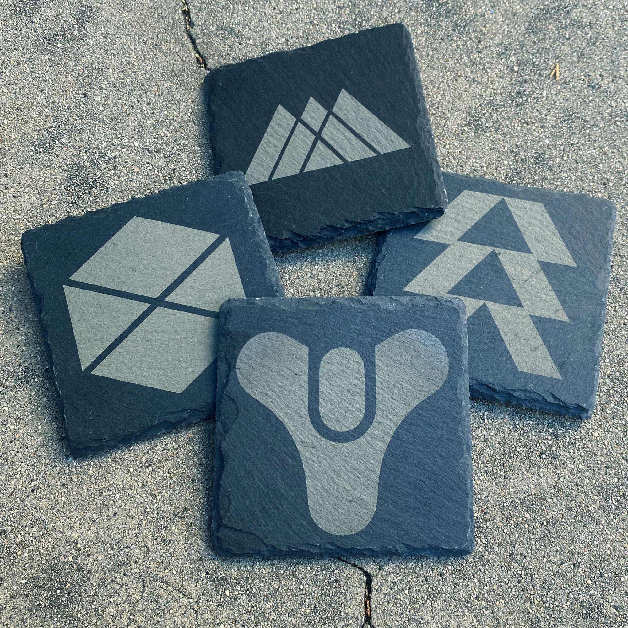 Four dark grey coasters made from slate against a concrete background. Each one has a Destiny logo engraved onto it.