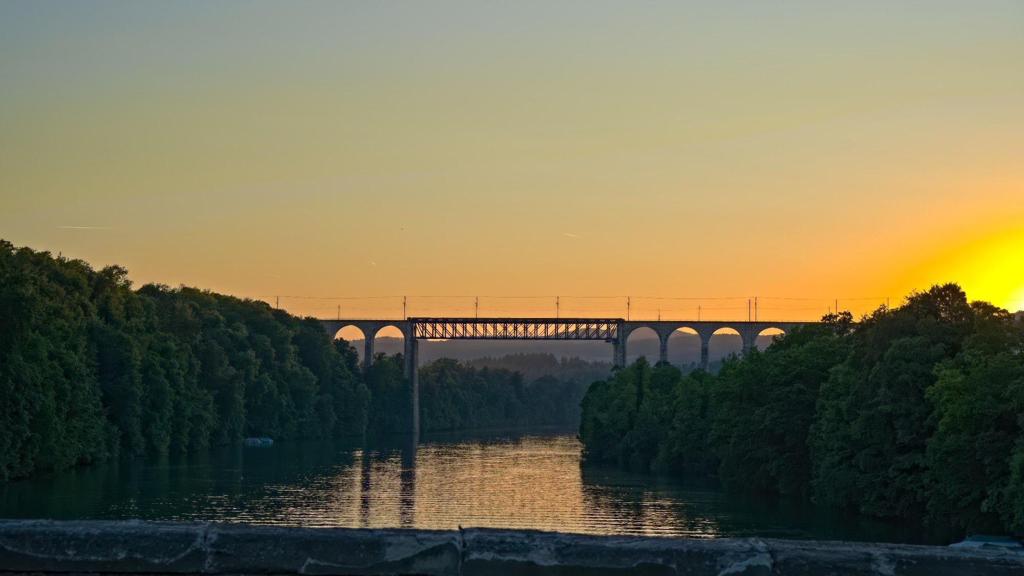 Viadukt für Eisenbahnen über einen Fluss=Rhein bei Sonnenuntergang. Wolkenlos, orangfarbener Himmel mit Verlauf ins Blau.
Der Rhein ist auf beiden Seiten dicht mit Bäumen bewachsen.