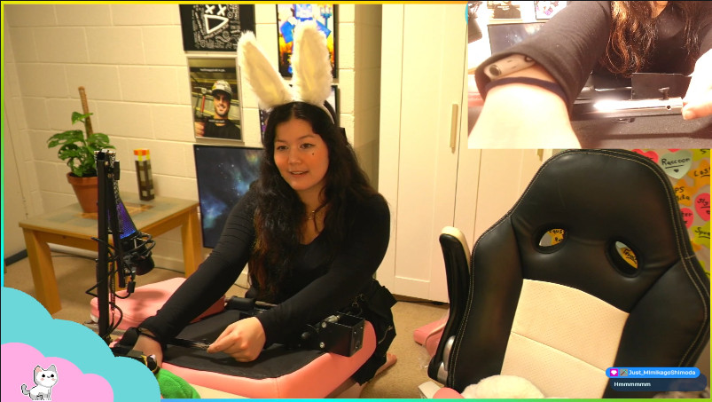 A slightly blurred picture from AngelNaomi's chair-building stream. She is sitting on the ground in a full black outfit, with her arms straight out holding chair parts. The parts are black and pink, and it looks like the underside of the chair. There is another chair, her usual chair, on the rightside, and a separate camera on the top right side of the screen. AngelNaomi is wearing cute long bunny ears in this shot.