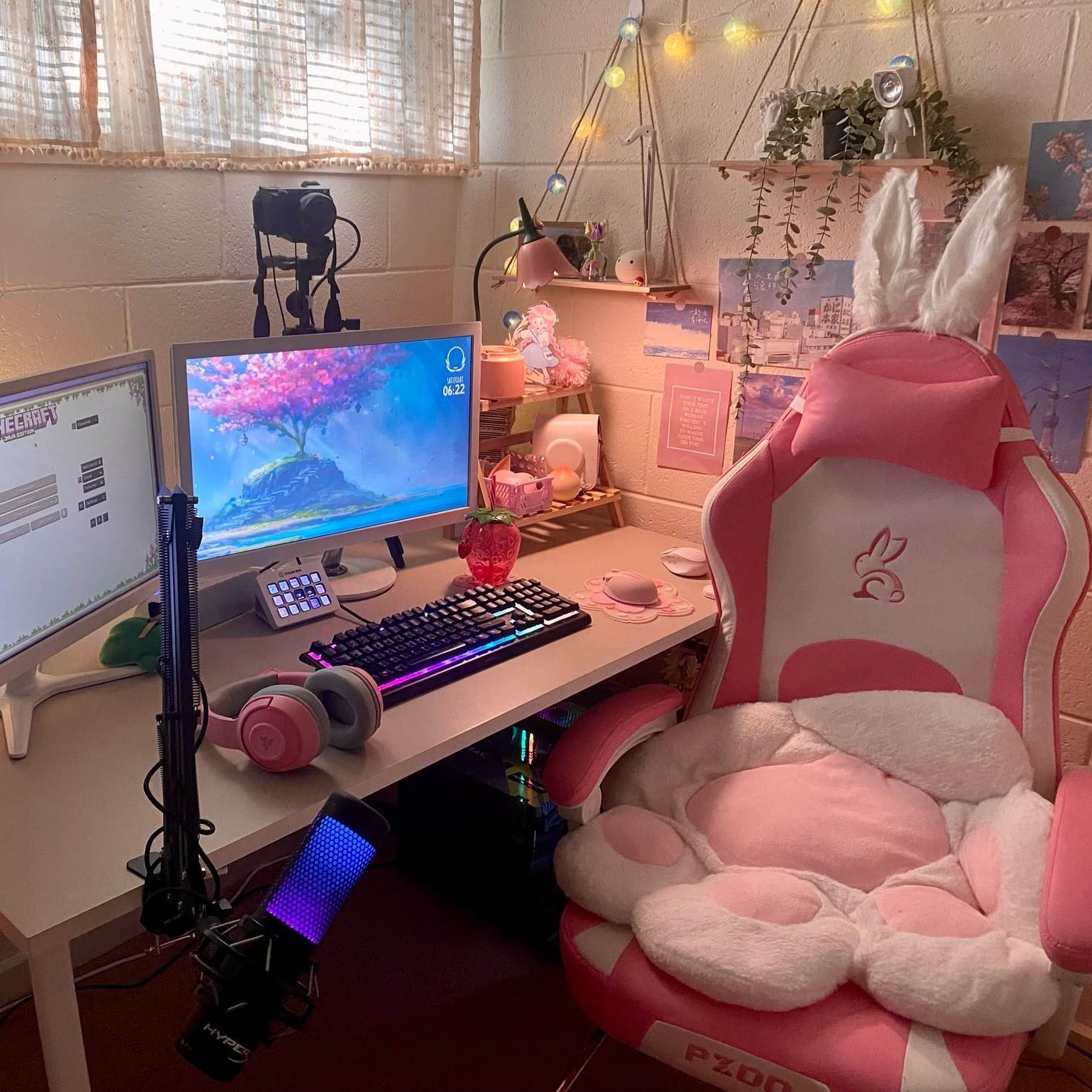 AngelNaomi's desk setup. It's very pink! There are 2 white-framed monitors, 1 black keyboard, and a large pink and white chair. The chair takes up about half of the frame. There is white bunny ears on top of the chair, with a paw print pillow sitting on the chair. The chair also has a logo of a rabbit. In the background is misc desk items, including a lamp, camera, drinkbottle, headphones, and mouse. On the wall behind there is a couple shelves with small items on it, plus some images in the background printed out and stuck to the wall. Behind the monitors there is also a camera.